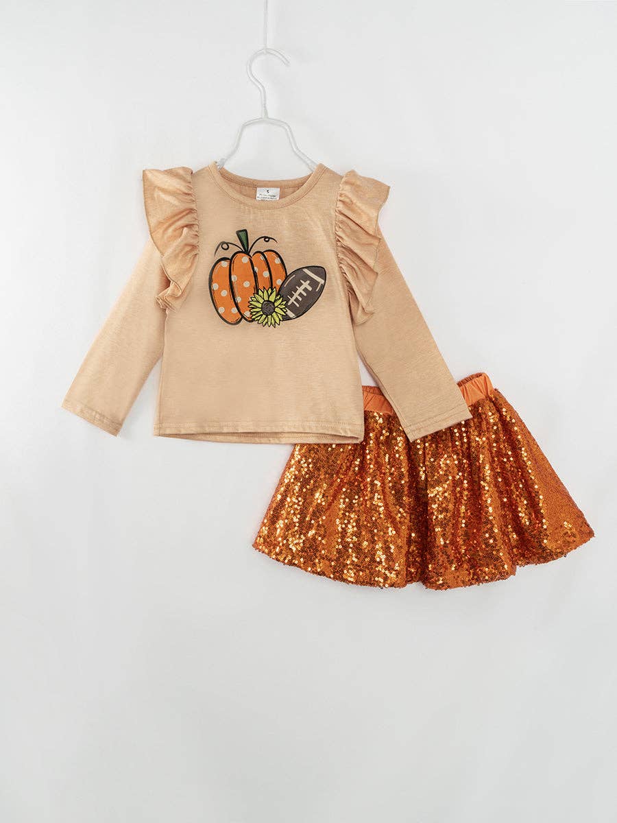 A festive toddler girl's fall outfit featuring a beige long-sleeve top with ruffled shoulders. The shirt is adorned with a graphic of an orange polka-dotted pumpkin, a football, and a sunflower. Paired with a shimmering orange sequin skirt, this ensemble is perfect for autumn-themed events or Thanksgiving celebrations. The playful combination of textures and seasonal motifs makes it both stylish and fun.