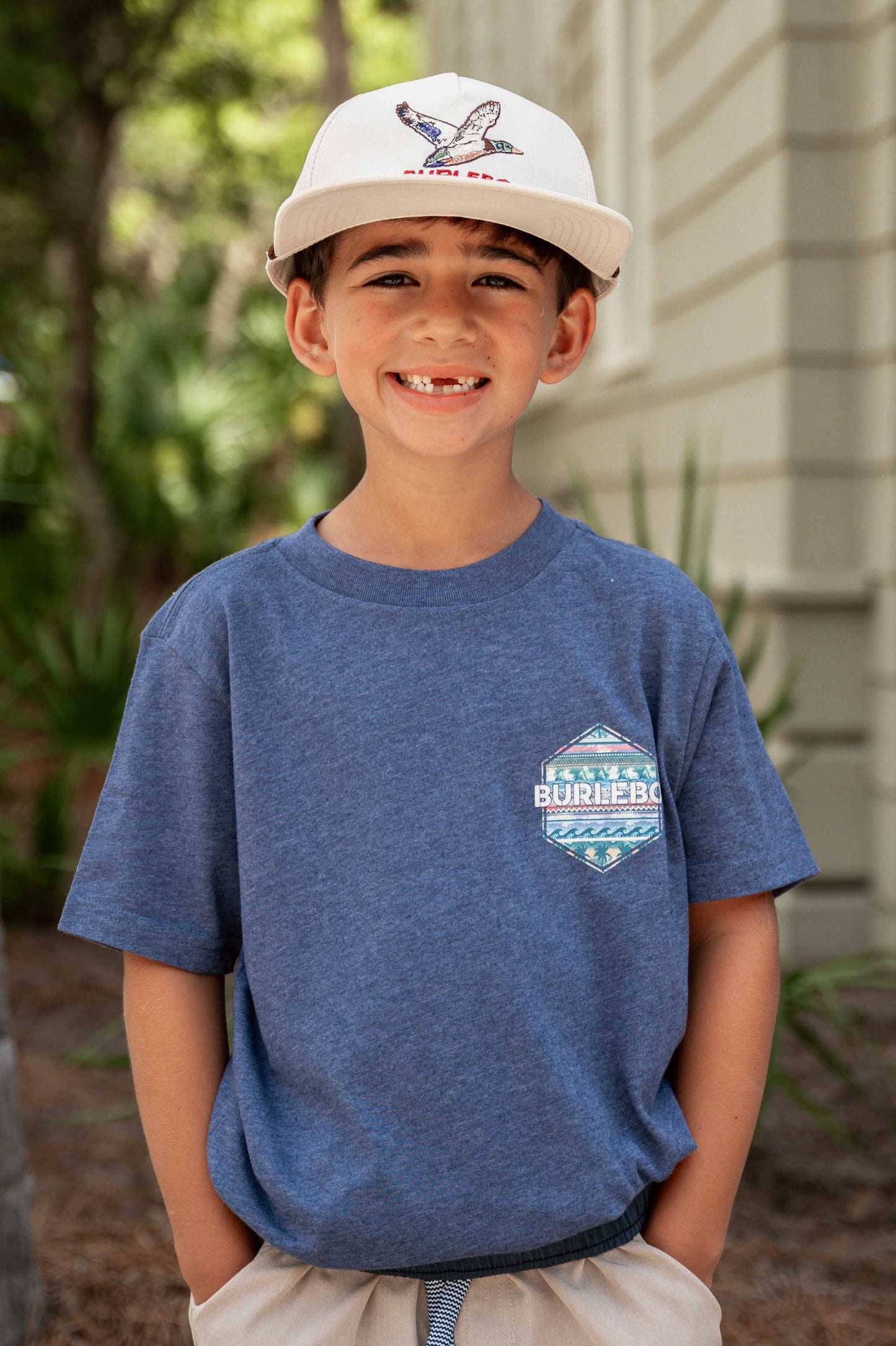 A young boy wearing a light beige backward cap and a blue short-sleeve t-shirt with a colorful geometric print design on the back. The design features tropical patterns with palm trees, ocean waves, and tribal-inspired elements, along with the text "BURLEBO" in bold white letters and "Find Your Wild" below it. He pairs the shirt with light beige shorts, standing outdoors near lush greenery and a tree trunk. The relaxed, nature-inspired outfit evokes a casual, adventurous vibe.