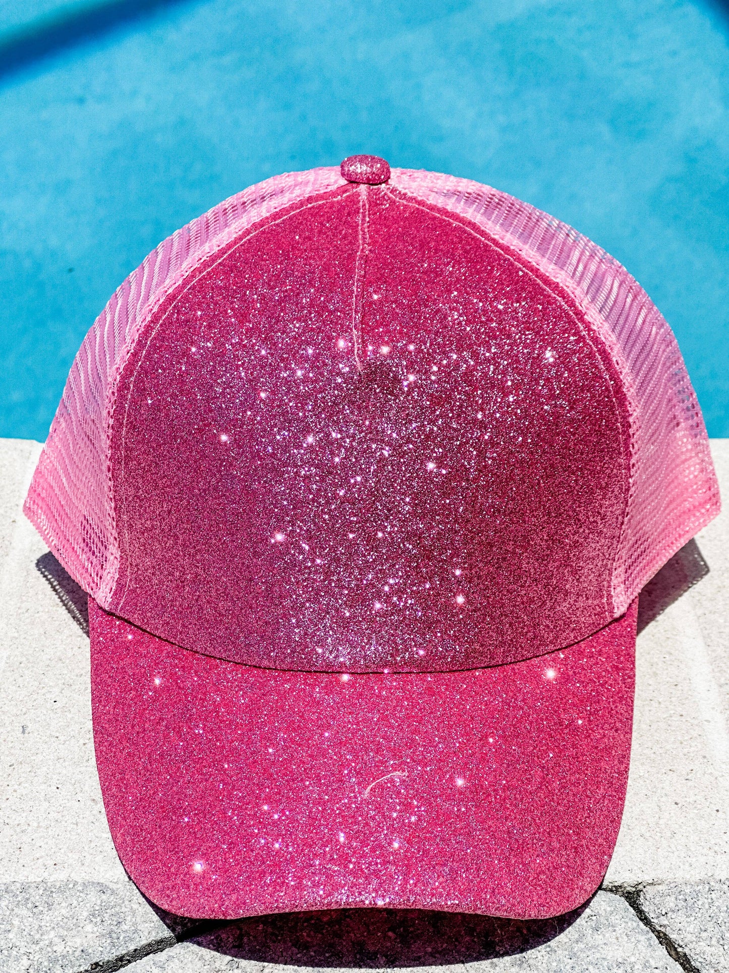"Pink glitter trucker hat with a mesh back, displayed in front of a bright blue pool background, shown from both the front and back views."