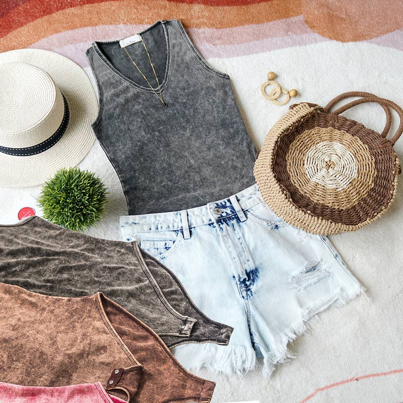 Flat lay of a boho summer outfit with a gray acid-wash tank top, light blue distressed denim shorts, a straw bag, a straw hat, and other tank tops in earthy tones arranged alongside.