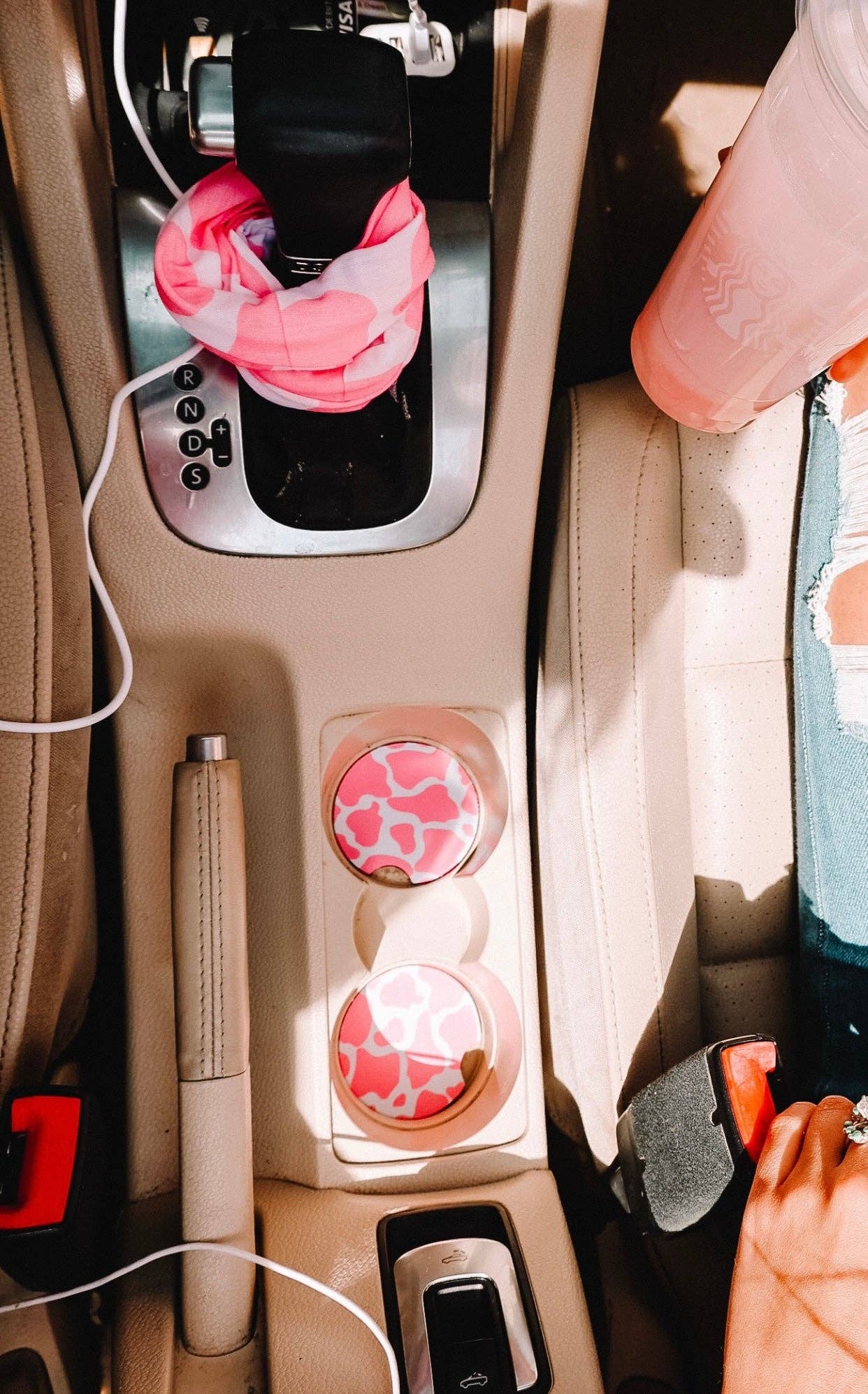 "Car interior featuring a beige console with pink cow-print cup holder coasters, a pink patterned scrunchie on the gear shift, and a matching pink drink tumbler on the passenger seat."