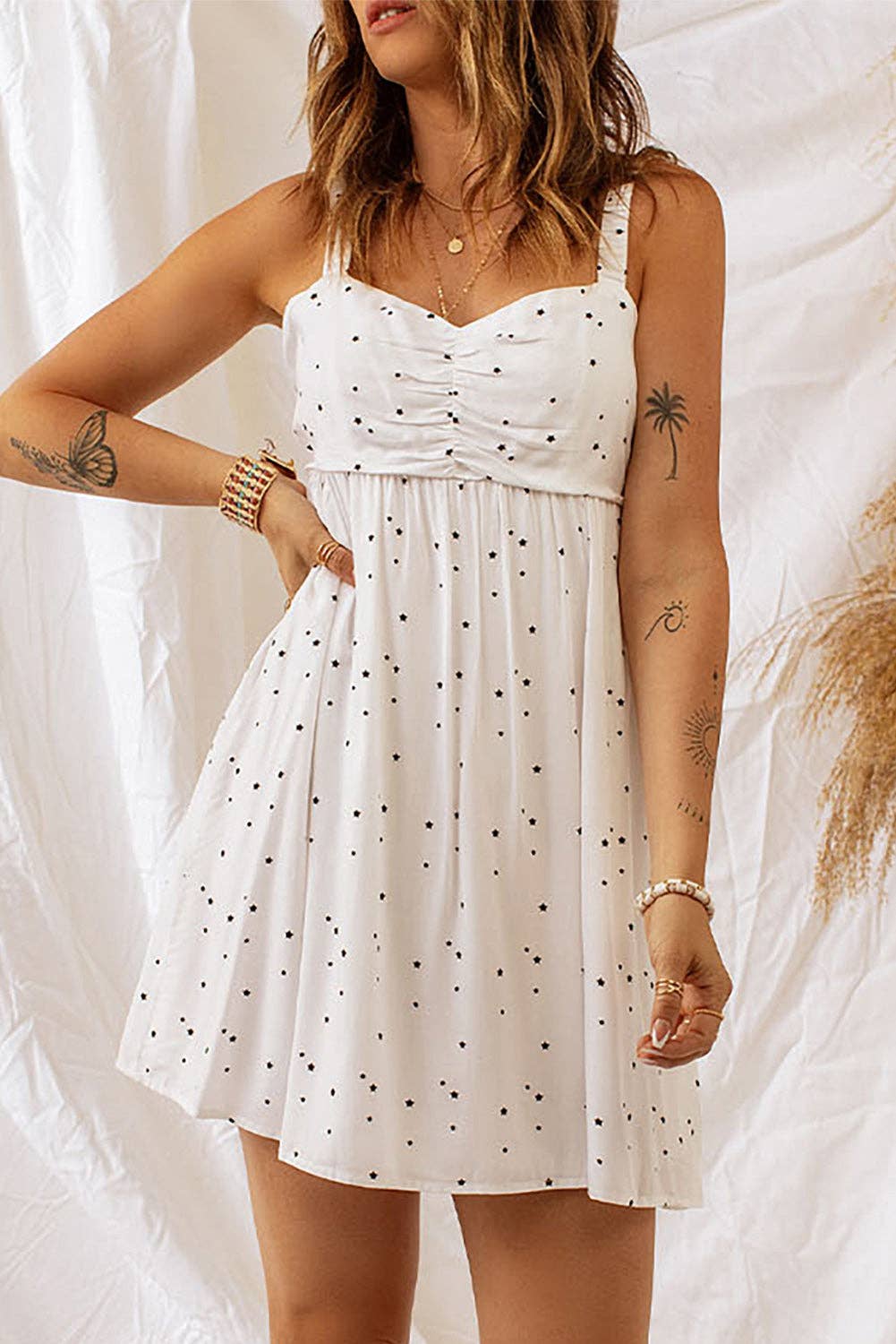 A woman wearing a white sundress with thin straps, ruched detailing at the bust, and a black polka dot pattern throughout. The dress flares out slightly, giving it a flowy look. She accessorizes with layered gold necklaces, a bracelet, and has tattoos on her arm, including a palm tree design. The background features soft white fabric and dried grasses, adding a bohemian vibe.