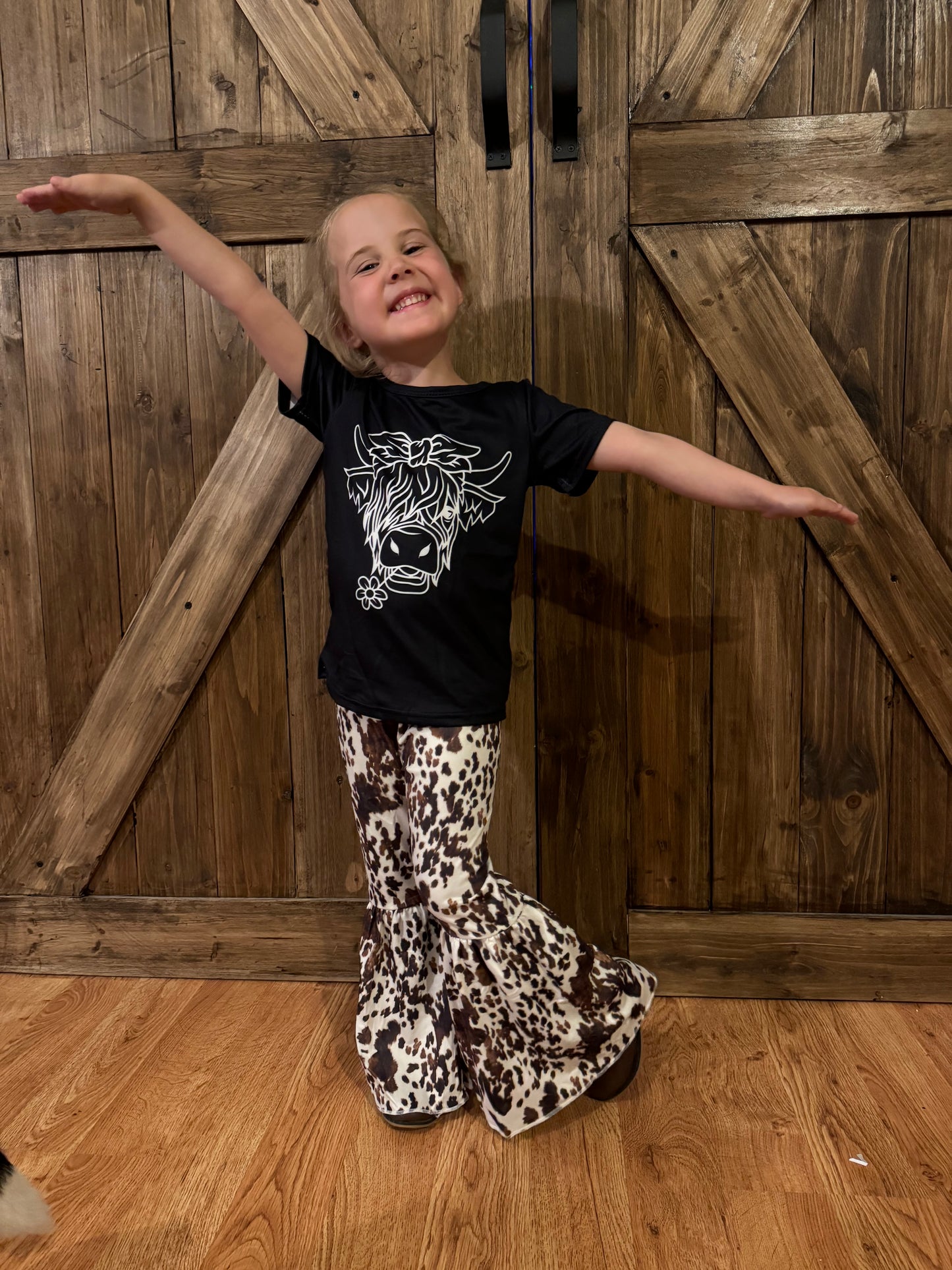 Cheerful young girl posing with arms outstretched, wearing a black T-shirt featuring a white graphic of a Highland cow with a bow and flower detail. She pairs it with flared bell-bottom pants in a bold brown-and-white cow print pattern. The rustic wooden barn door background and warm-toned flooring add a country-inspired charm to the scene.
