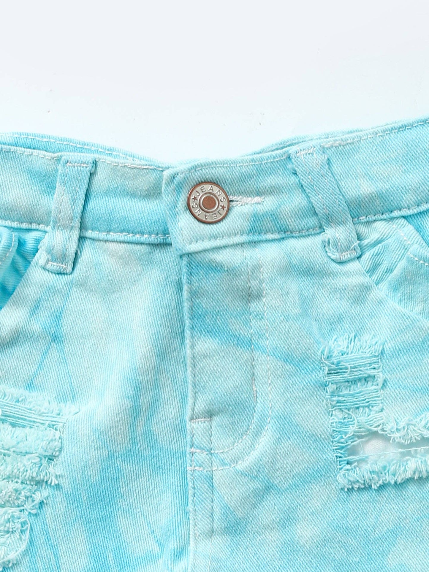Adorable light blue tie-dye denim shorts for girls, featuring distressed detailing, subtle ruffle accents along the sides, and a button closure. Styled with a sleeveless white button-front top, these shorts create a playful, summery outfit perfect for casual outings or photoshoots. Modeled in front of a green floral backdrop with neon 'Bomb' signage.