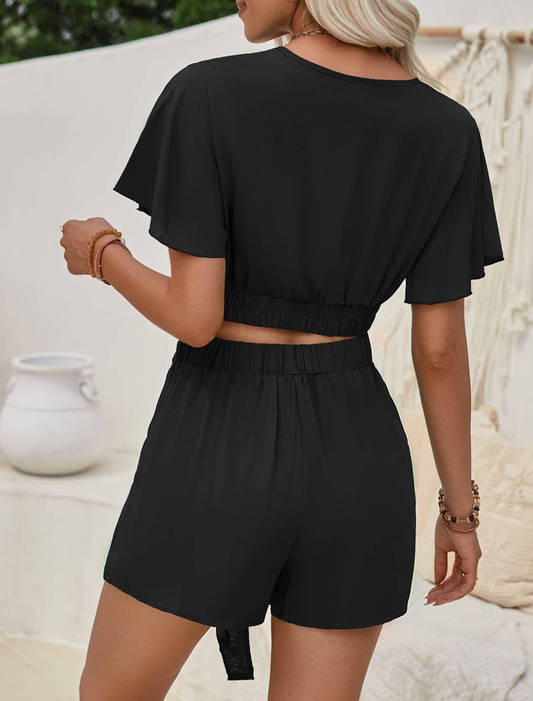 A woman models a chic black two-piece set featuring a wrap-style crop top with flutter sleeves and a matching high-waisted skirt with a front tie detail. She accessorizes with layered gold necklaces, hoop earrings, a mix of gold and beaded bracelets, and a gold statement ring. Her wavy blonde hair complements the neutral-toned boho background, which includes macrame wall hangings, textured cushions, and a white ceramic vase. The outfit exudes an elegant, relaxed summer vibe.