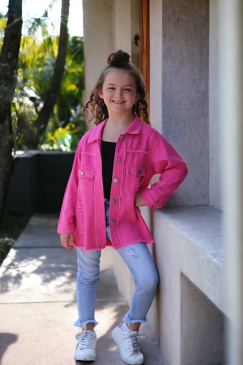 Smiling young girl wearing a bright pink distressed shacket over a black top, paired with light-wash denim jeans. Styled outdoors with a modern architectural backdrop, this outfit is a perfect mix of bold and casual fashion for kids.