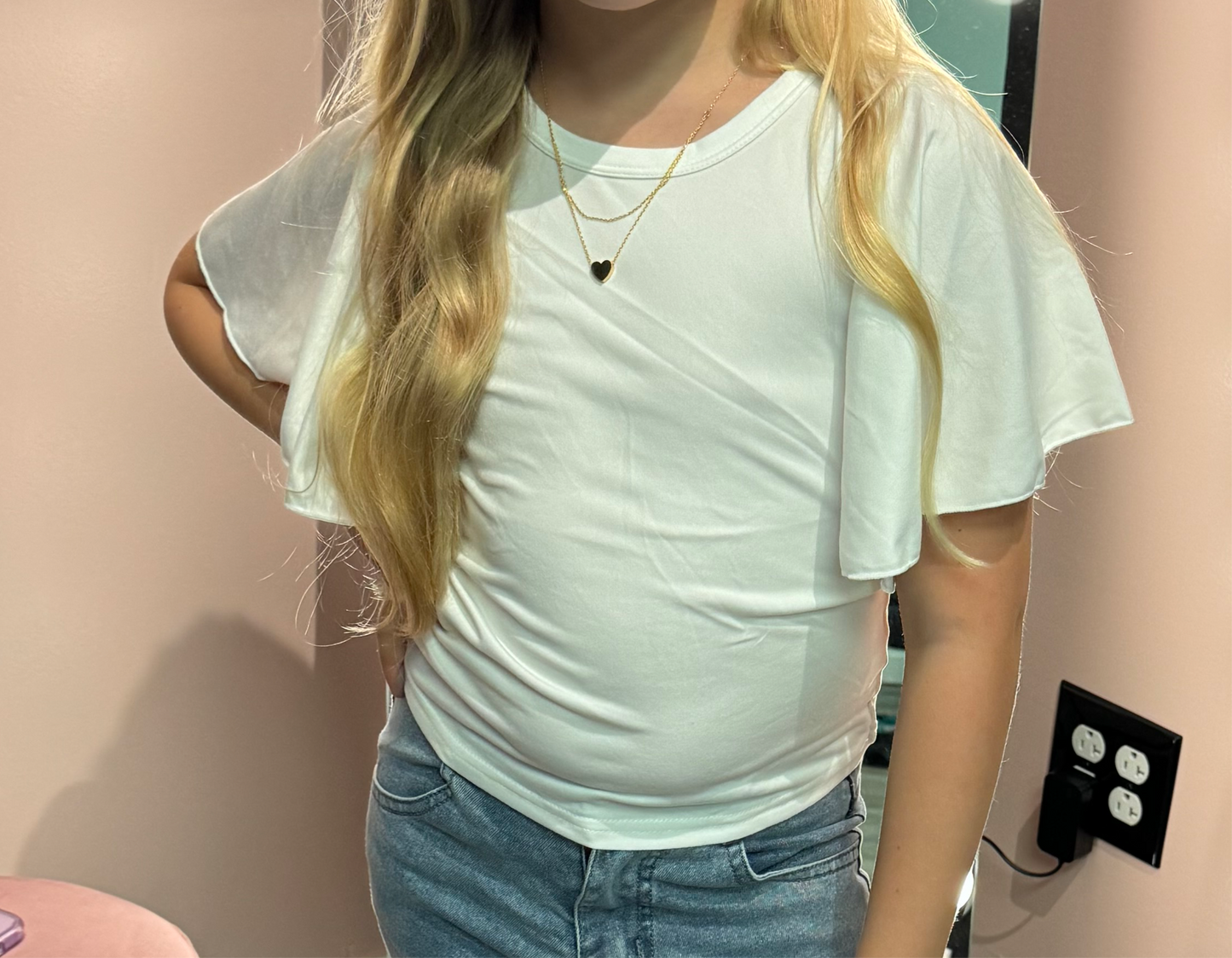 Close-up of a young girl’s torso wearing a white flutter-sleeve top paired with layered gold necklaces, one with a small heart pendant. The focus is on the outfit and accessories.
