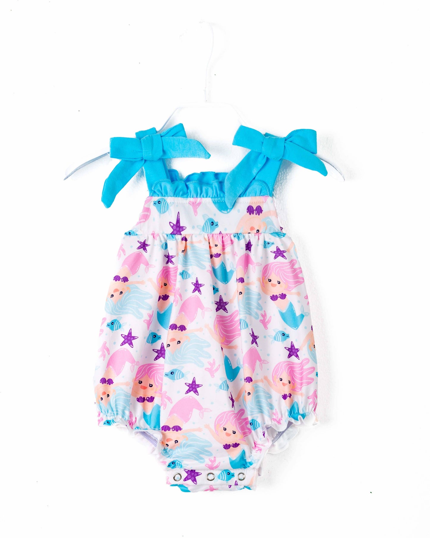A colorful baby romper featuring a whimsical underwater mermaid theme. The fabric is adorned with pink and blue mermaids, starfish, fish, seashells, and coral motifs against a white background. The romper has bright blue straps tied into playful bows at the shoulders, adding a charming touch. The bottom has snap closures for easy diaper changes, and the gathered elastic at the legs ensures a comfortable fit. Displayed on a white hanger, the romper is perfect for summer or themed photoshoots.