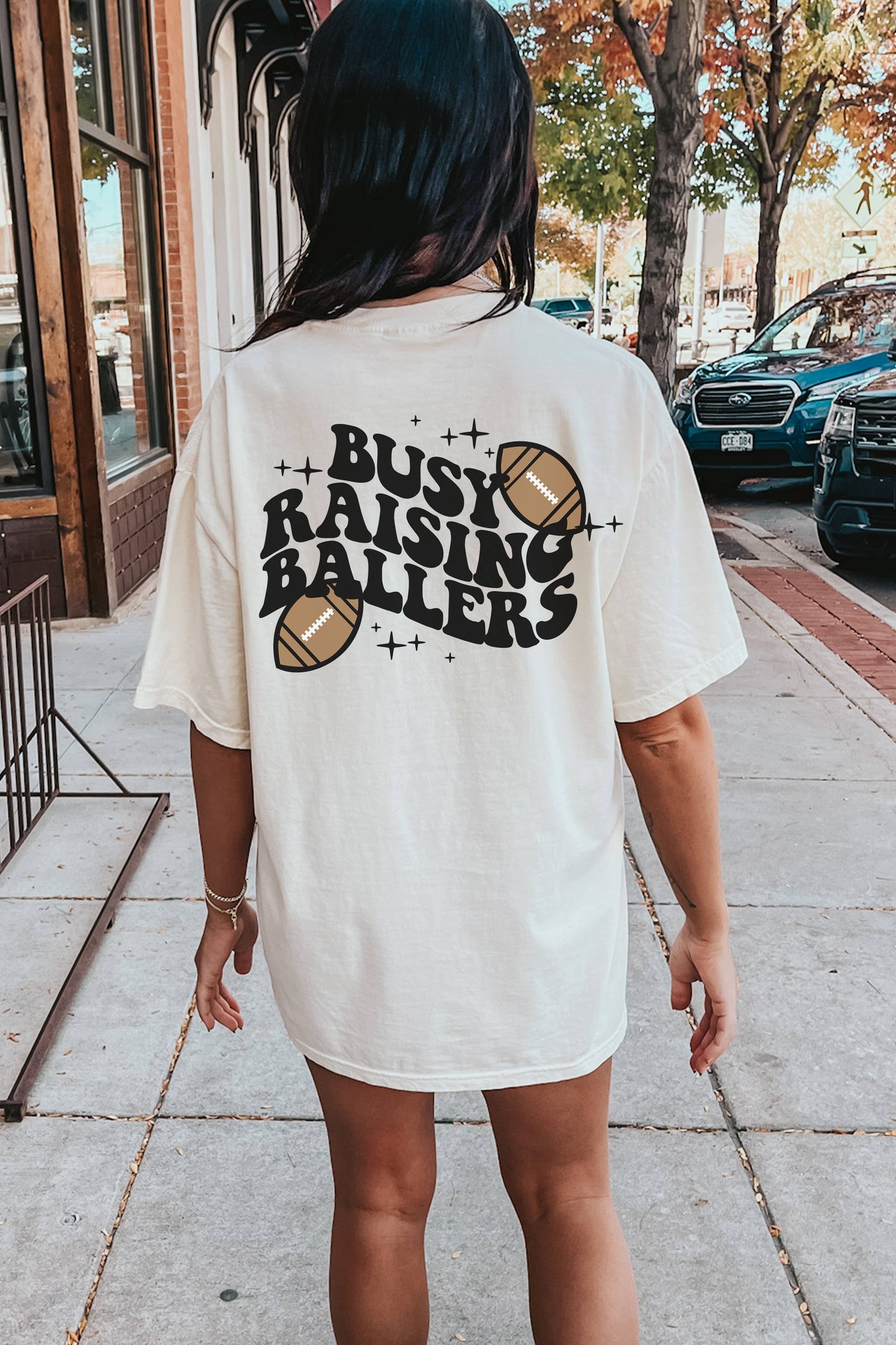 The back of the shirt showcases bold black text reading "BUSY RAISING BALLERS" surrounded by two football graphics and sparkles. The playful design emphasizes a sporty, family-oriented theme, making it perfect for football moms or sports enthusiasts. The casual backdrop of a sidewalk with parked cars and fall foliage enhances the relaxed, everyday appeal of the outfit.