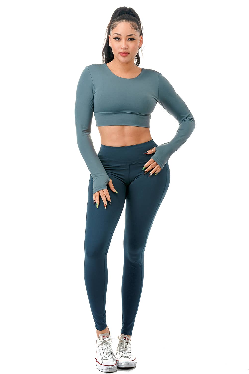A model wearing a stylish teal two-piece activewear set consisting of a long-sleeve cropped top and high-waisted leggings. The top features a fitted design with thumbholes and a crisscross strap detail at the open back, providing both support and style. The leggings have a smooth, high-rise waistband with a seamless look, complementing the fitted silhouette. The model completes the outfit with white sneakers and a high ponytail, showcasing a sporty and chic appearance. Perfect for workouts, yoga,