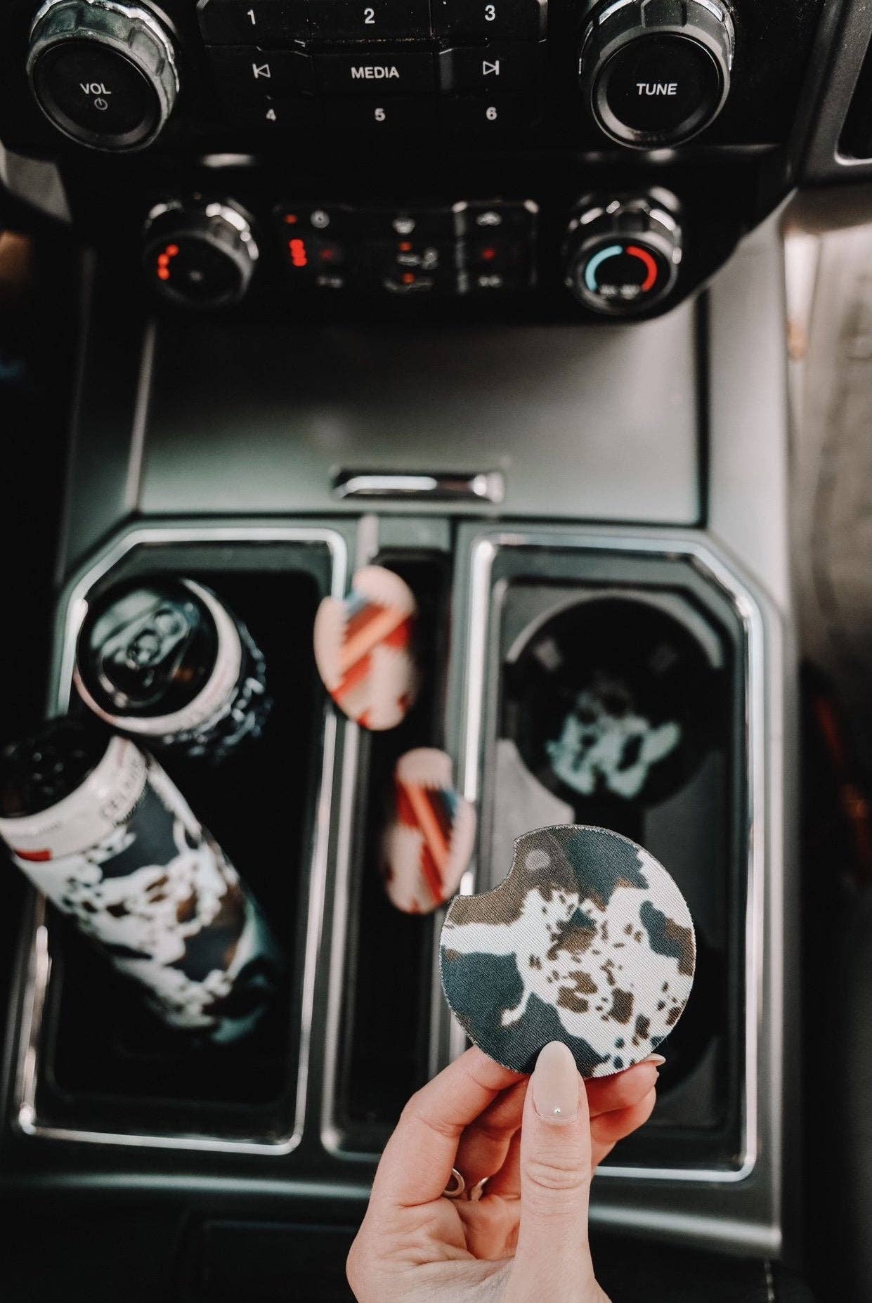 Cow-print car coasters shown in a vehicle's cup holder, with a close-up of one being held. The dashboard features modern controls, emphasizing the coasters' stylish and functional design for Midwestern-inspired auto accessories.
