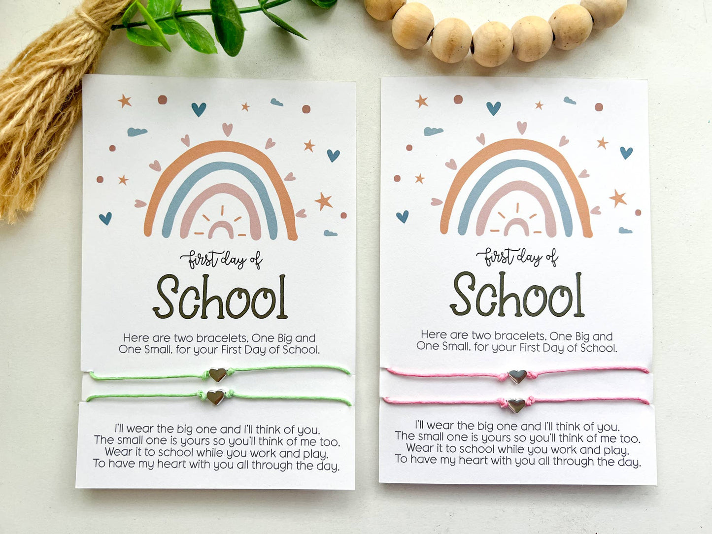 Two "First Day of School" cards featuring a pastel rainbow design with hearts, stars, and clouds. Each card includes a pair of matching string bracelets—one larger and one smaller—with heart-shaped charms in the center. The cards read: "Here are two bracelets. One Big and One Small, for your First Day of School." Below, a heartfelt message explains the bracelets' purpose for connection during the day. One card has mint green bracelets, and the other has pink. Both are placed on a white surface.