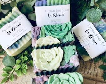 An arrangement of "In Bloom" eucalyptus-scented soap bars with colorful green, lavender, and cream swirls, showcasing floral-shaped tops and intricate designs. The bars are displayed among lush greenery, highlighting their nature-inspired aesthetic and handcrafted appeal.