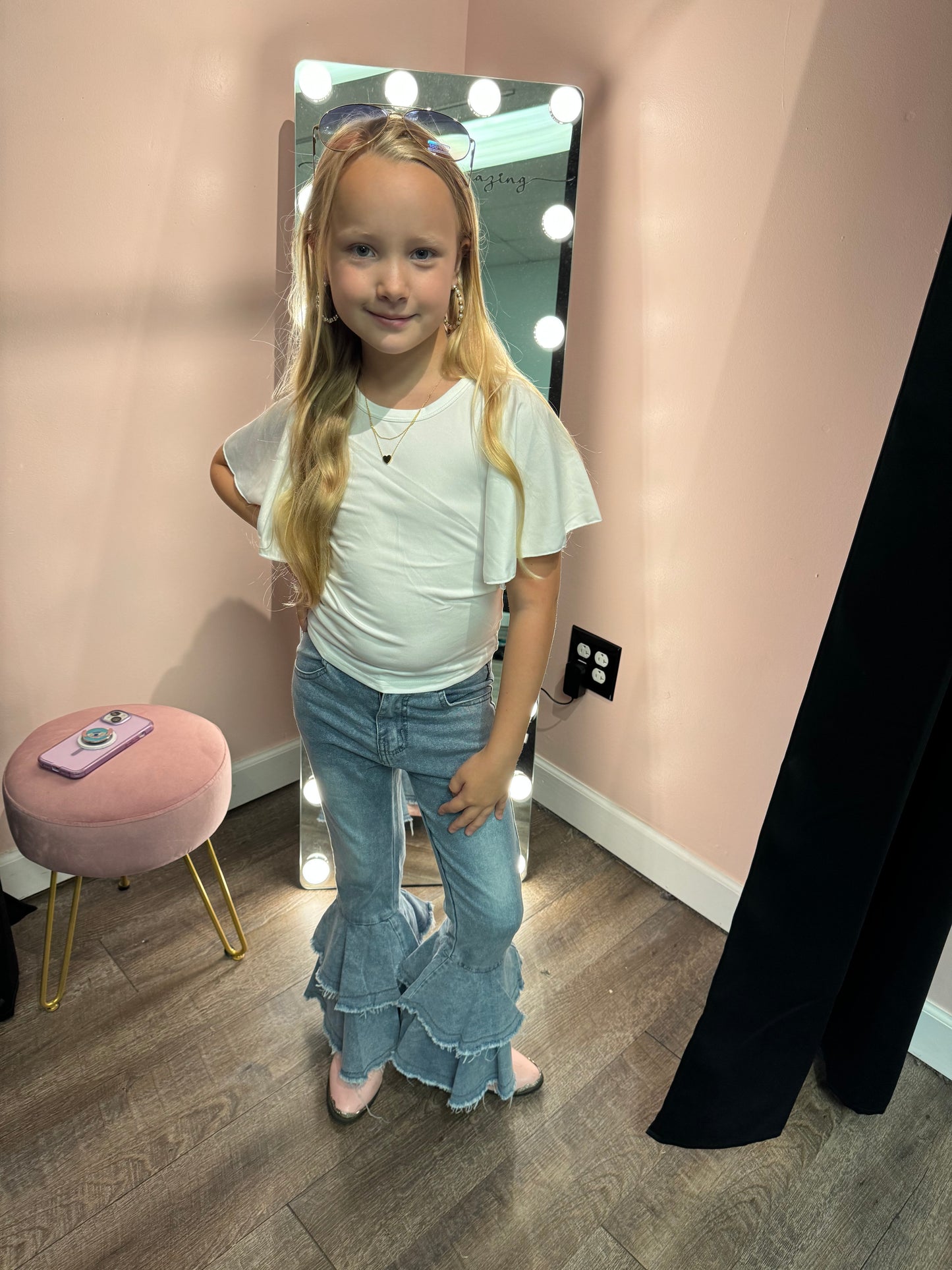 A young girl posing confidently in light blue tiered denim bell-bottom jeans and a white flutter-sleeve top. She is accessorized with hoop earrings, layered necklaces, and pink shoes, standing in front of a makeup mirror surrounded by lights.