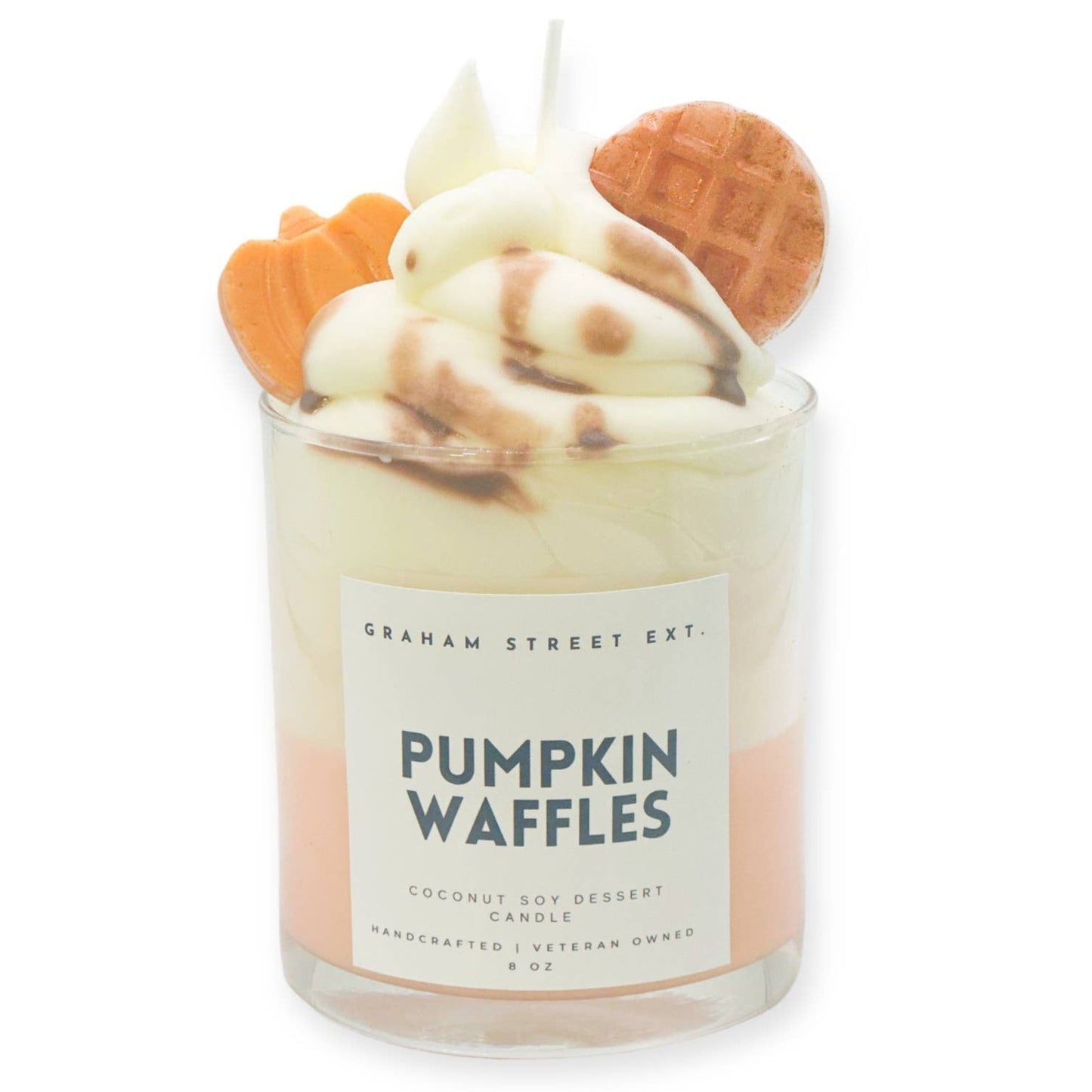A decorative "Pumpkin Waffles" scented candle by Graham Street Ext., crafted to resemble a dessert. The candle is made of coconut soy wax and features a whipped cream-like top swirled with brown accents mimicking syrup. It is embellished with realistic wax shapes, including a miniature waffle and a pumpkin, for a festive autumn theme. The glass container displays a minimalistic label with the text "Pumpkin Waffles," highlighting its handcrafted and veteran-owned origins. Designed as an 8 oz. candle, it exud