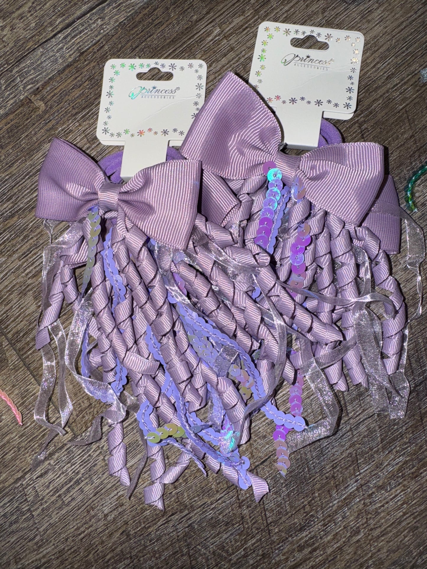 A set of purple hair bows with cascading curly ribbons and sparkly sequin details, displayed on branded packaging labeled "Princess Accessories." The bows are placed on a wooden surface, showcasing their whimsical and elegant design.
