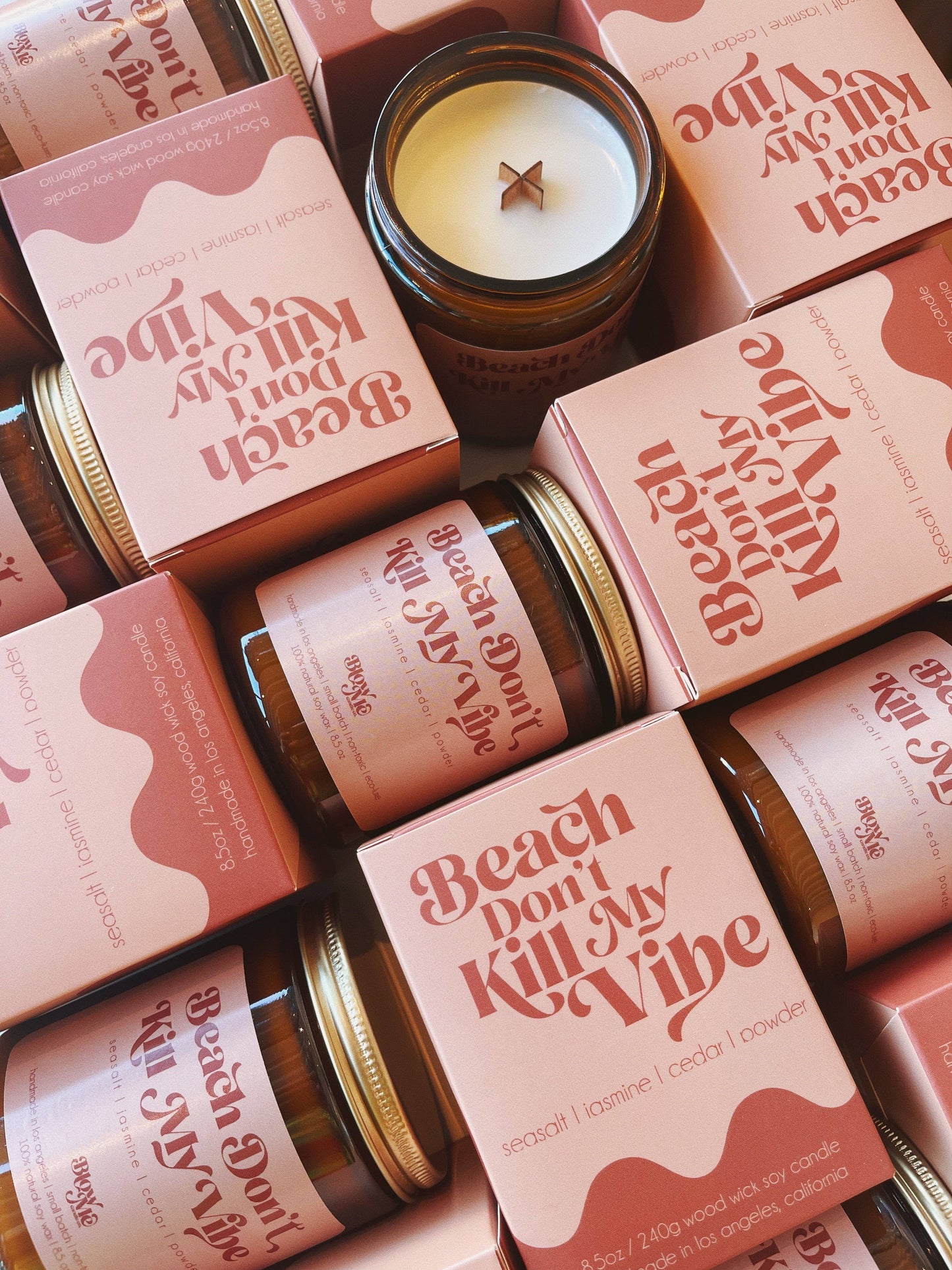 Flat-lay display of multiple soy candles by Blow Me Candle Co., with pink retro-themed packaging labeled 'Beach Don’t Kill My Vibe.' Each box highlights the scent profile of seasalt, jasmine, cedar, and powder, alongside amber glass jars with wooden wicks. The arrangement exudes a chic and modern aesthetic, ideal for promoting artisanal home fragrances.