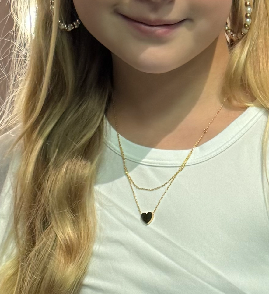 A close-up of a young girl wearing a double-layer gold heart necklace paired with a white top. The simple heart pendant hangs delicately, complemented by large pearl hoop earrings, showcasing the necklace's elegant and versatile style.