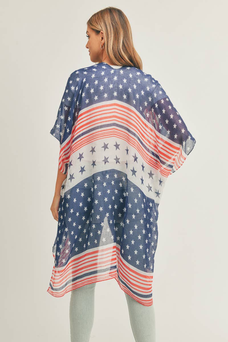A woman wearing a lightweight patriotic shawl with a design featuring blue with white stars and red and white stripes. The shawl drapes elegantly over her shoulders, complementing her white ribbed top. She has long, wavy blonde hair and is accessorized with gold hoop earrings, posing against a neutral background.
