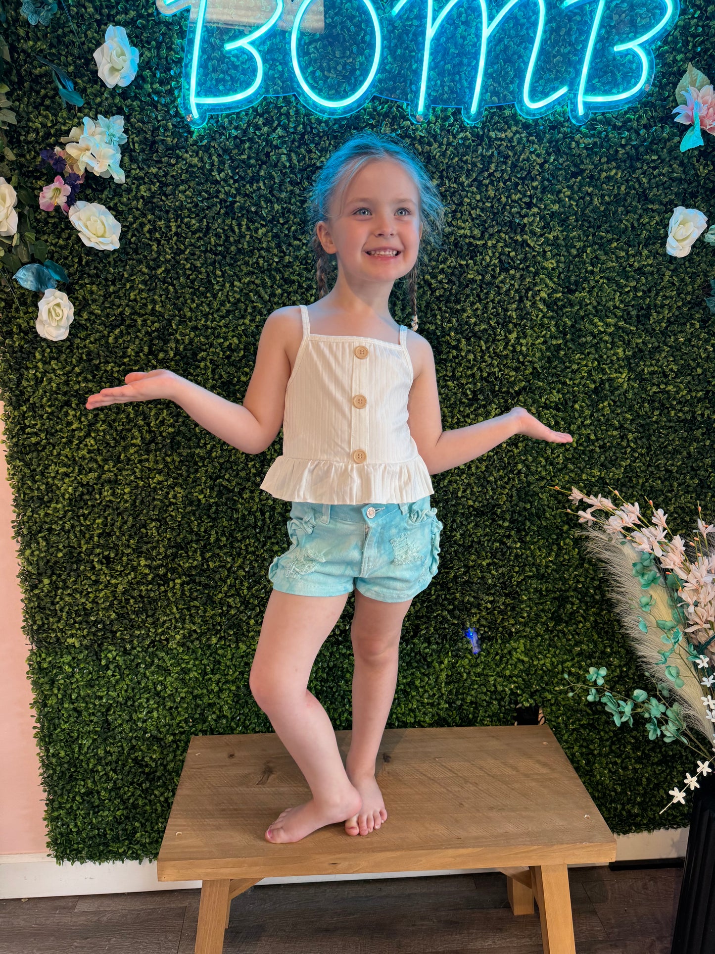 Adorable light blue tie-dye denim shorts for girls, featuring distressed detailing, subtle ruffle accents along the sides, and a button closure. Styled with a sleeveless white button-front top, these shorts create a playful, summery outfit perfect for casual outings or photoshoots. Modeled in front of a green floral backdrop with neon 'Bomb' signage.