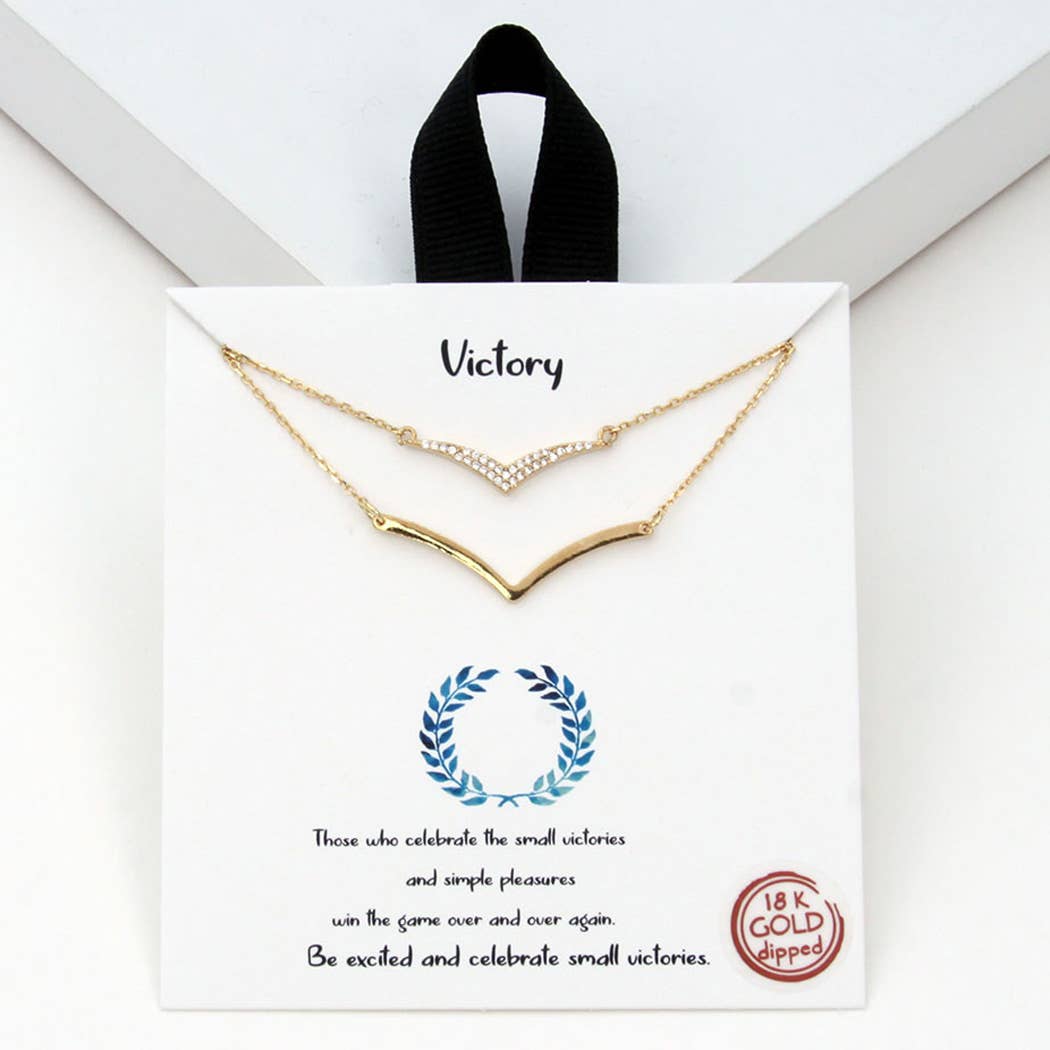 A gold double-layered necklace displayed on a white card with the word "Victory" written at the top. The top necklace features a delicate chevron design adorned with sparkling rhinestones, while the bottom layer has a smooth gold chevron pendant. The card includes an inspiring message: "Those who celebrate the small victories and simple pleasures win the game over and over again. Be excited and celebrate small victories." It also highlights that the necklace is 18K gold-dipped, with a laurel wreath graphic