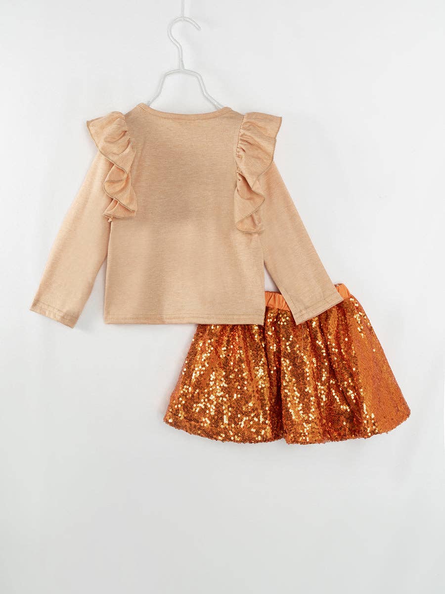 A festive toddler girl's fall outfit featuring a beige long-sleeve top with ruffled shoulders. The shirt is adorned with a graphic of an orange polka-dotted pumpkin, a football, and a sunflower. Paired with a shimmering orange sequin skirt, this ensemble is perfect for autumn-themed events or Thanksgiving celebrations. The playful combination of textures and seasonal motifs makes it both stylish and fun.