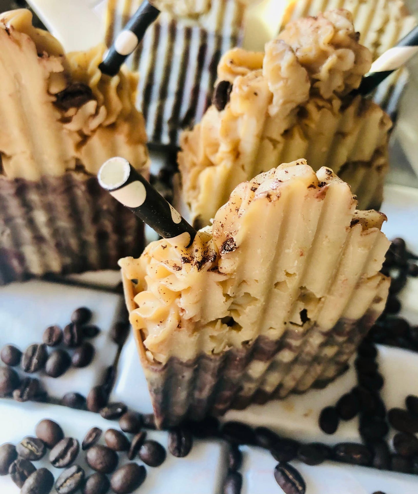 A close-up view of artisan soap bars designed to look like a frothy coffee drink, featuring a textured, creamy beige top resembling whipped cream and a rich brown base mimicking coffee layers. Each bar is adorned with a black-and-white striped straw and sprinkled with coffee grounds, surrounded by scattered coffee beans for a visually appealing, coffee-themed aesthetic.