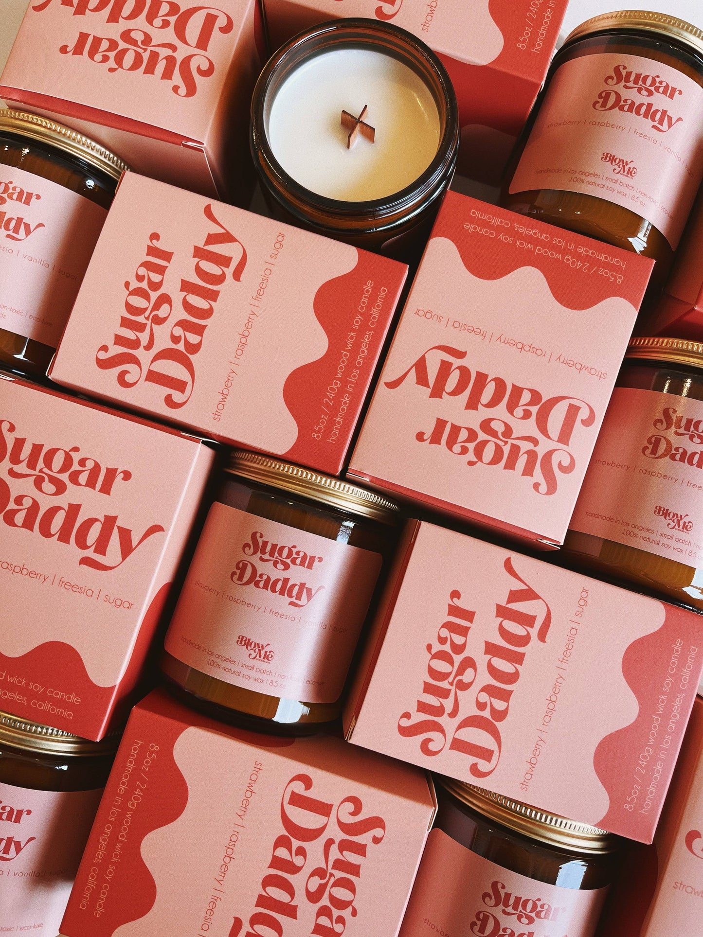 A collection of "Sugar Daddy" candles and their pink packaging stacked together. The packaging and labels highlight the candle's scent profile, featuring strawberry, raspberry, freesia, and sugar. One candle is open, showing a wooden wick, emphasizing the artisanal design.