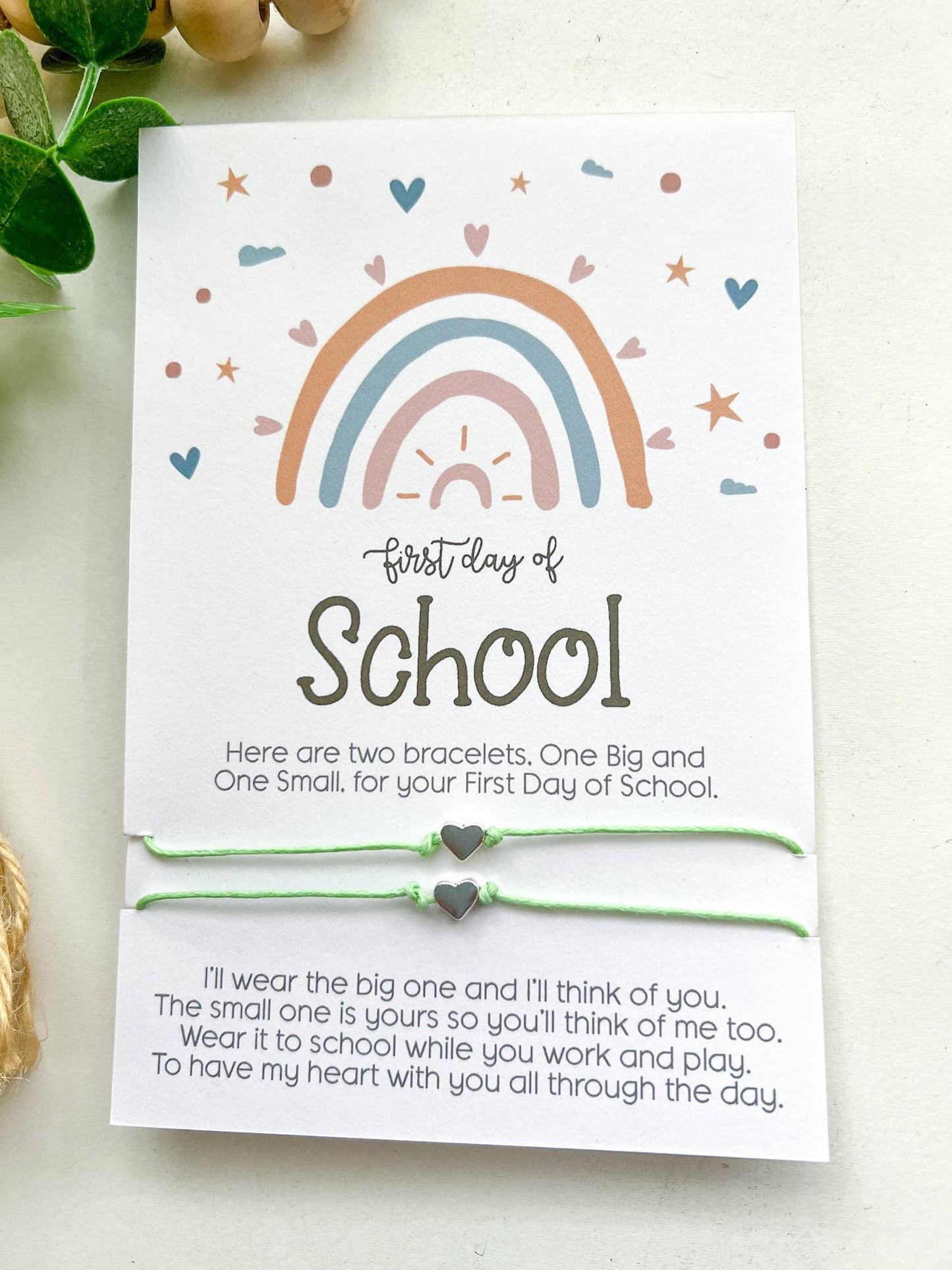 Two "First Day of School" cards featuring a pastel rainbow design with hearts, stars, and clouds. Each card includes a pair of matching string bracelets—one larger and one smaller—with heart-shaped charms in the center. The cards read: "Here are two bracelets. One Big and One Small, for your First Day of School." Below, a heartfelt message explains the bracelets' purpose for connection during the day. One card has mint green bracelets, and the other has pink. Both are placed on a white surface.
