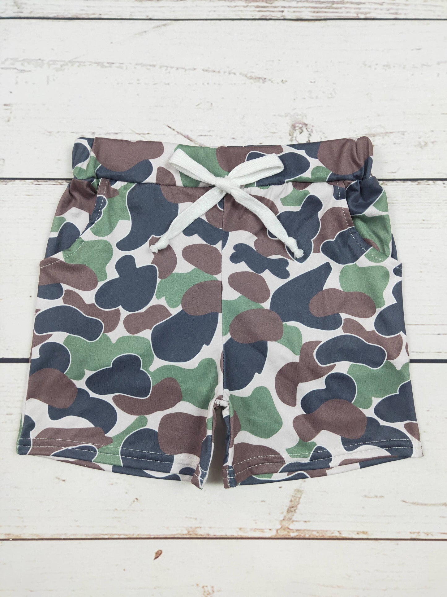 "Kids' camo-print shorts with a green, brown, and navy pattern, featuring an adjustable white drawstring and side pockets, displayed on a rustic wooden background."