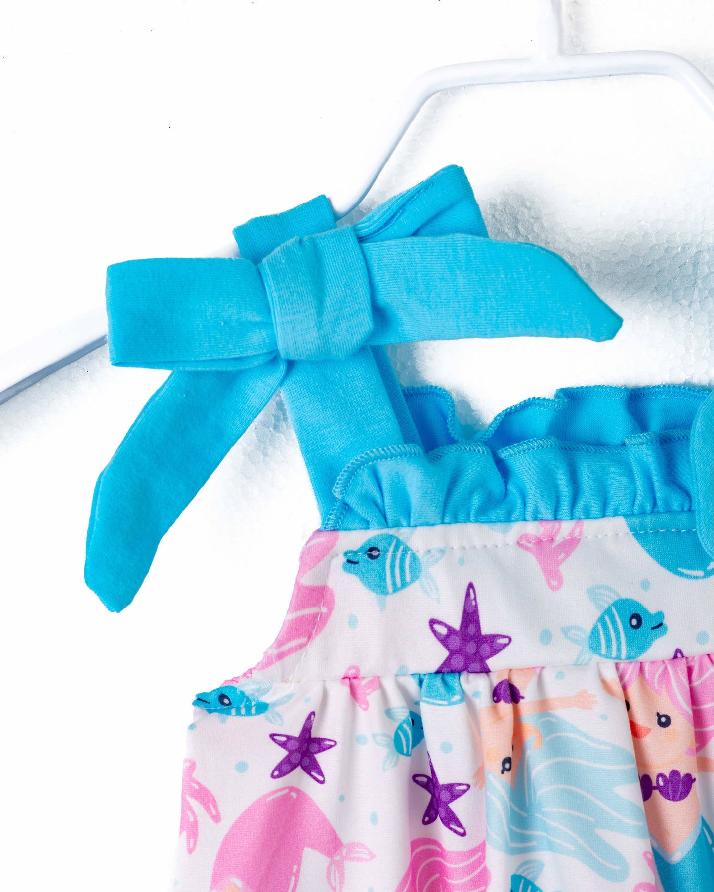 A colorful baby romper featuring a whimsical underwater mermaid theme. The fabric is adorned with pink and blue mermaids, starfish, fish, seashells, and coral motifs against a white background. The romper has bright blue straps tied into playful bows at the shoulders, adding a charming touch. The bottom has snap closures for easy diaper changes, and the gathered elastic at the legs ensures a comfortable fit. Displayed on a white hanger, the romper is perfect for summer or themed photoshoots.