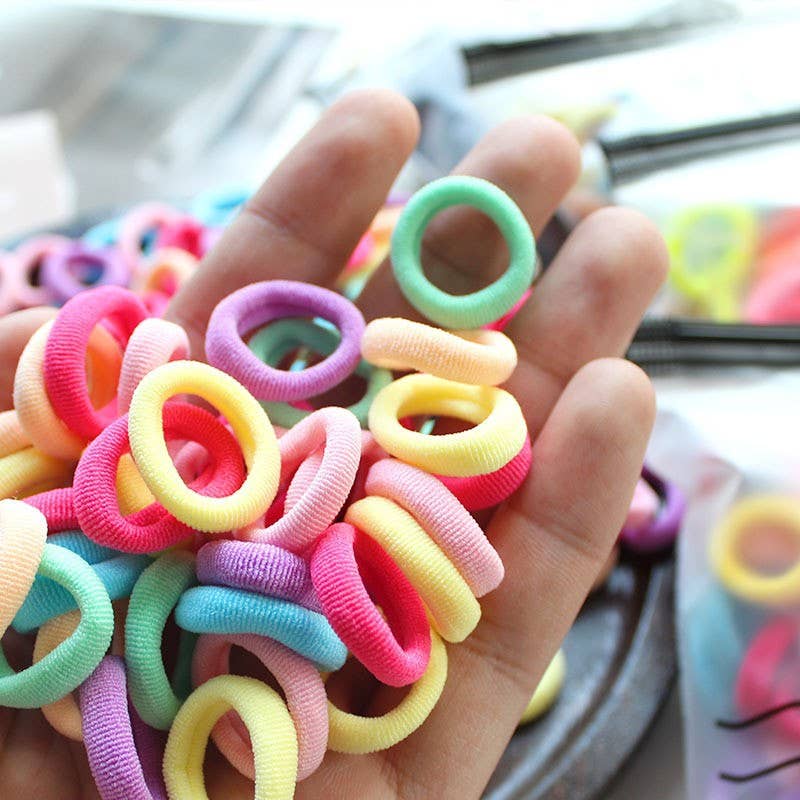 A small plastic zippered bag labeled "I Can Swim" is open, spilling out a colorful assortment of hair ties onto a white surface. The hair ties come in soft pastel shades, including pink, purple, yellow, green, and blue, creating a vibrant and playful display.