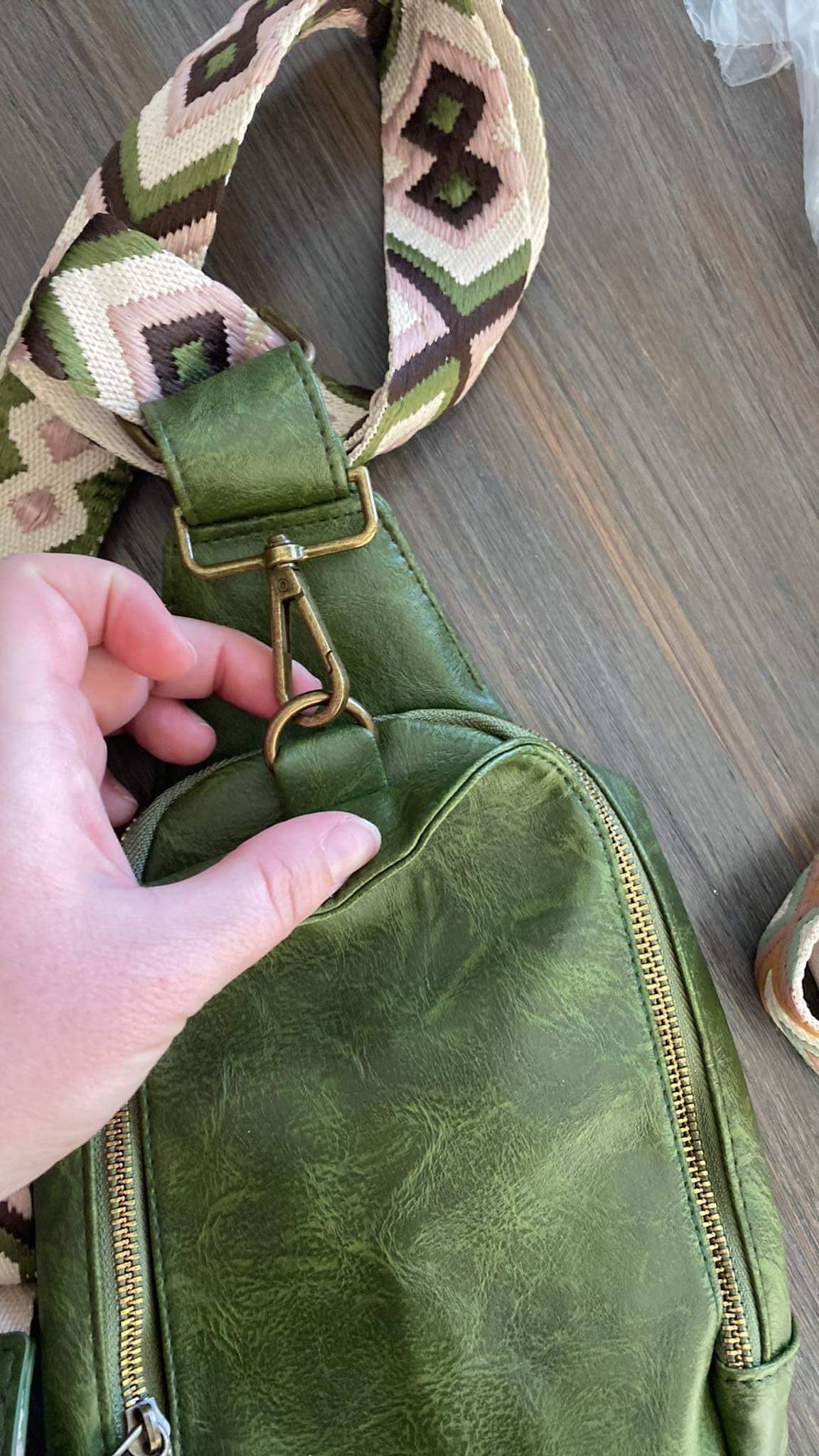 Alt text: A beige faux-leather sling bag with a zipper closure and a patterned strap featuring a tan and white zigzag diamond design. The bag is displayed on a dark wooden surface, showcasing its minimalist and stylish design.