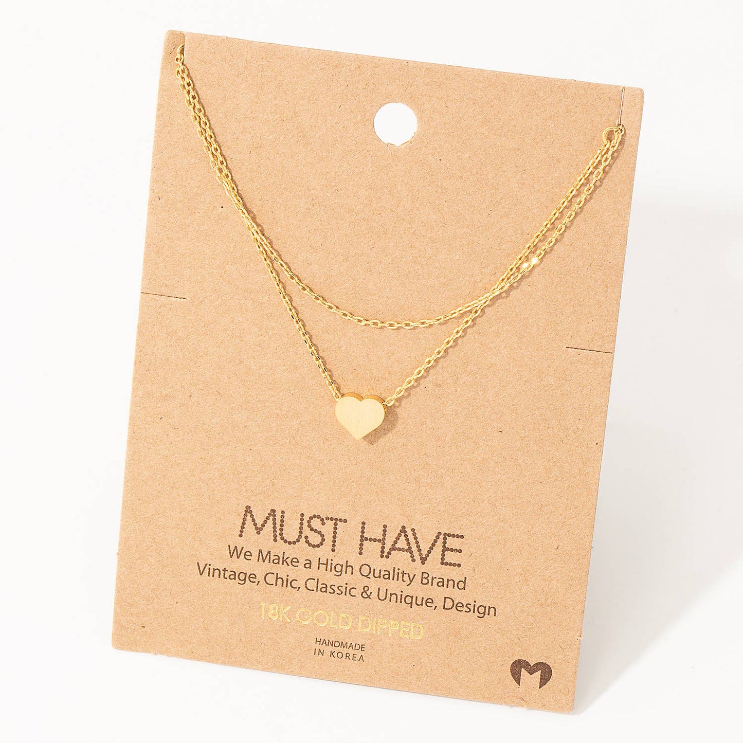 Minimalist gold heart pendant necklace displayed on a kraft cardstock backing labeled "MUST HAVE." The necklace features a delicate double-layer gold chain and a small heart charm, crafted with 18K gold-dipped material. Packaging highlights its vintage, chic, and handmade design, emphasizing quality craftsmanship.