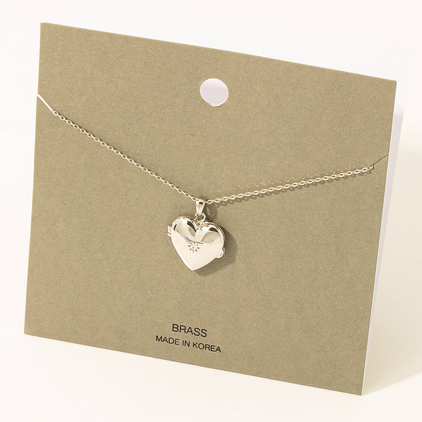 A delicate silver-tone heart locket necklace displayed on an olive-green card. The polished heart-shaped pendant features a subtle engraved starburst detail, adding a touch of elegance. The card includes the text "BRASS" and "Made in Korea," indicating the material and origin. A timeless and sentimental accessory perfect for gifting or personal wear.