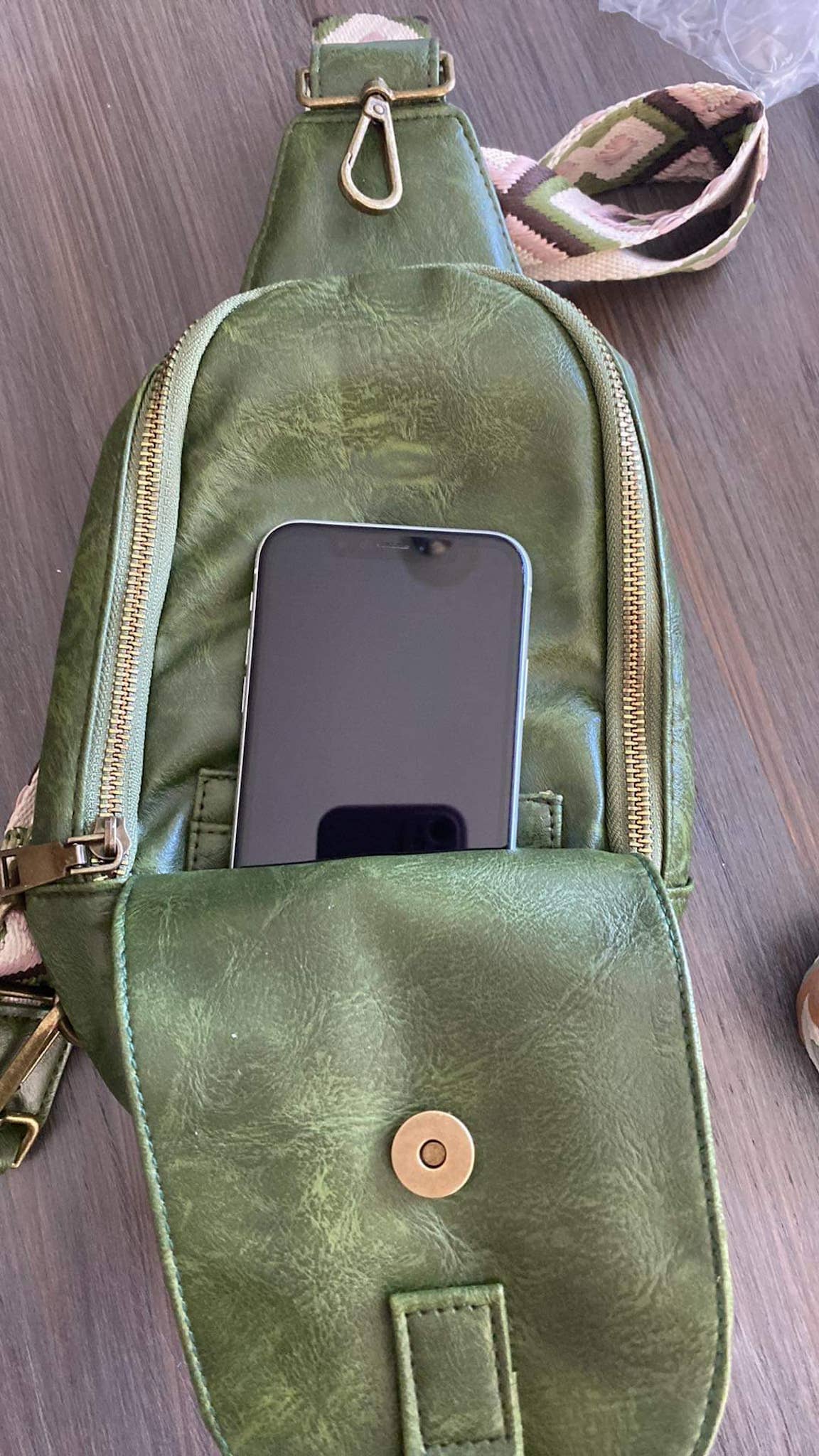 Alt text: A beige faux-leather sling bag with a zipper closure and a patterned strap featuring a tan and white zigzag diamond design. The bag is displayed on a dark wooden surface, showcasing its minimalist and stylish design.