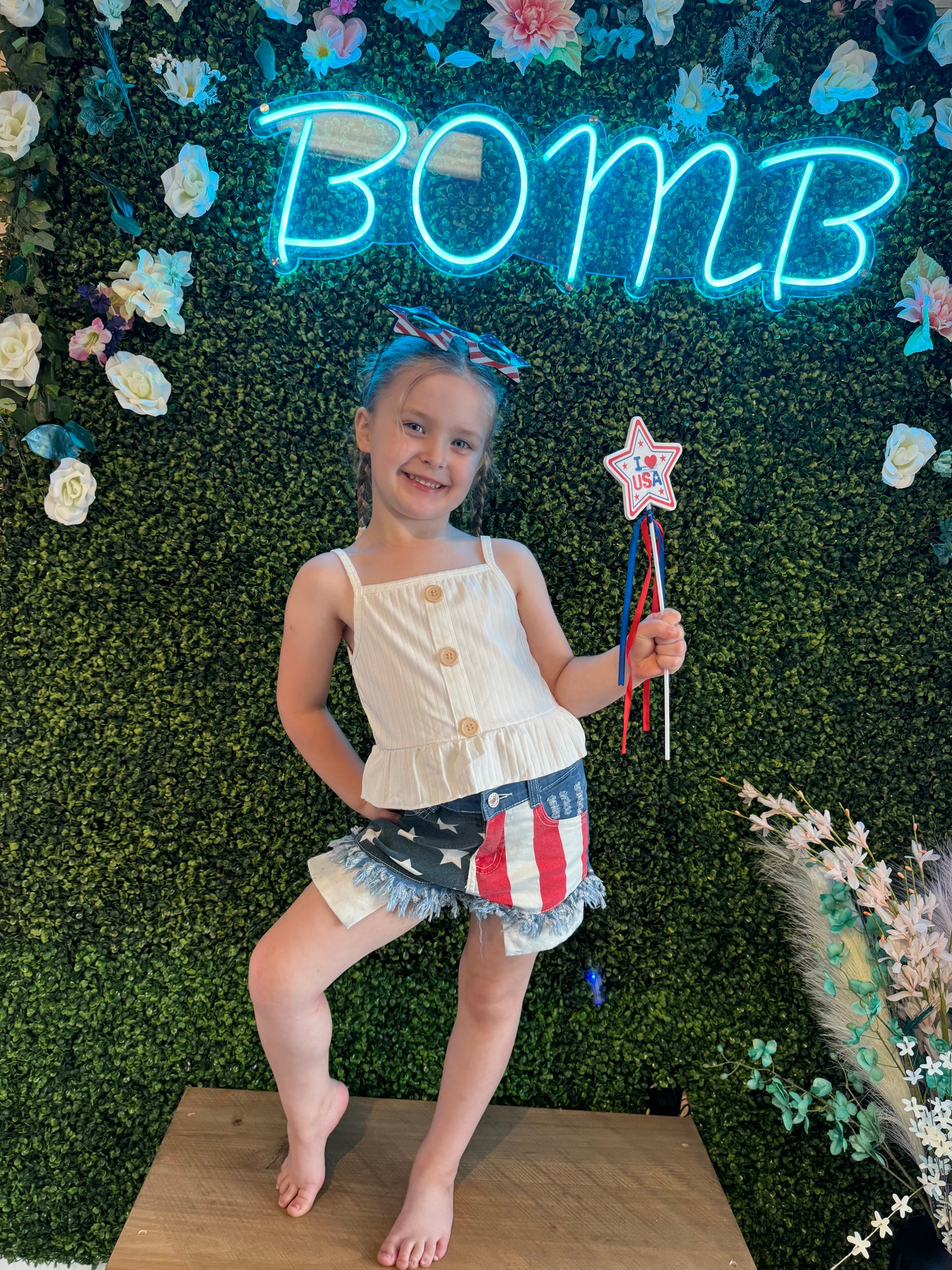 A young girl posing indoors in front of a green floral wall with a neon "BOMB" sign. She wears a white sleeveless button-up top with a ruffled hem and distressed denim shorts featuring a patriotic flag design. She accessorizes with a red, white, and blue bow in her hair and holds a star-shaped "I ❤️ USA" wand