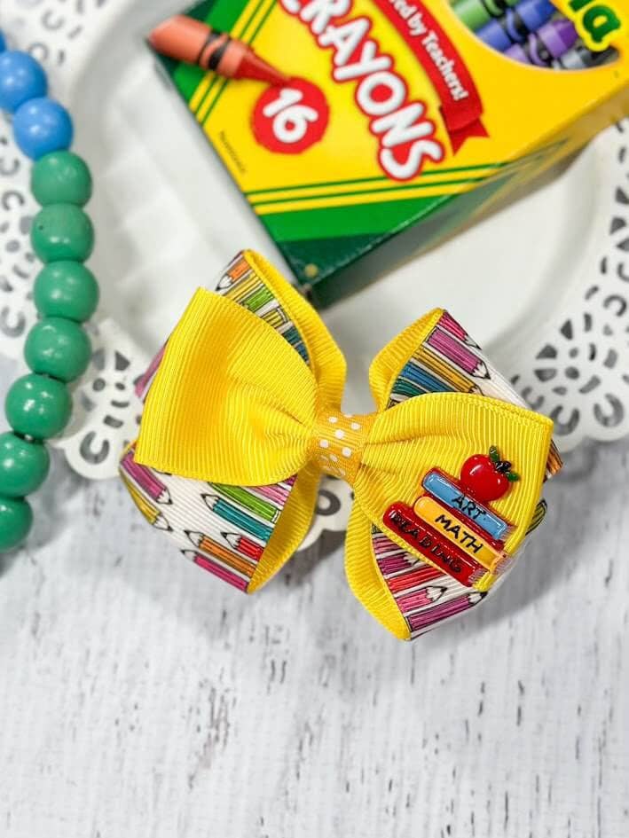 Alt text: A vibrant school-themed hair bow featuring a yellow ribbon layered with a patterned ribbon displaying colorful pencils. The bow is adorned with an enamel charm of stacked books labeled "Art," "Math," and "Reading," topped with a red apple. The bow is displayed on a decorative white plate, accompanied by a green beaded bracelet and a box of Crayola crayons in the background, adding to the back-to-school theme.