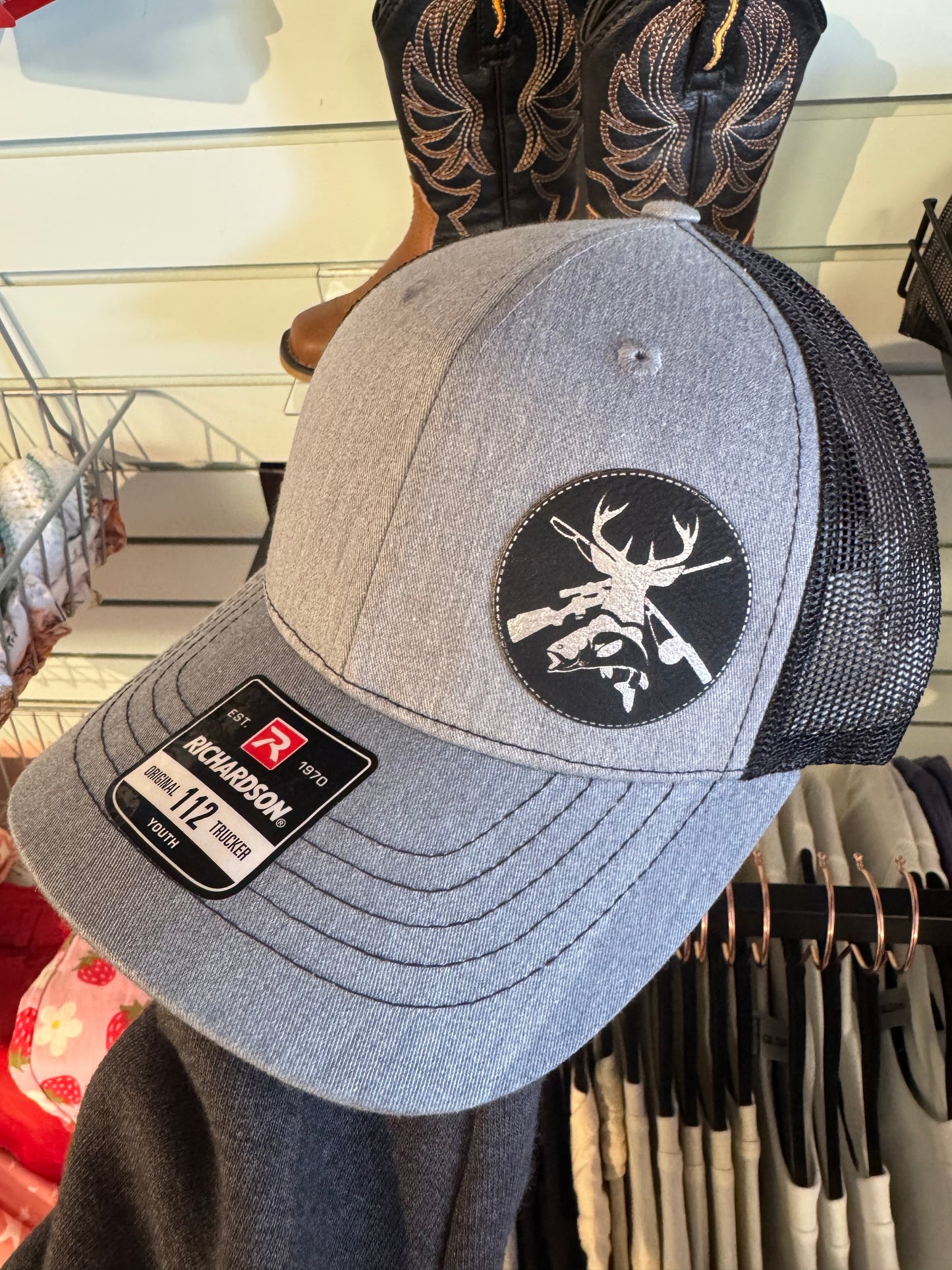 A gray and black trucker hat with a black mesh back and a circular patch on the front featuring a deer silhouette with crossed rifles. The hat is displayed on a retail rack with cowboy boots visible in the background.