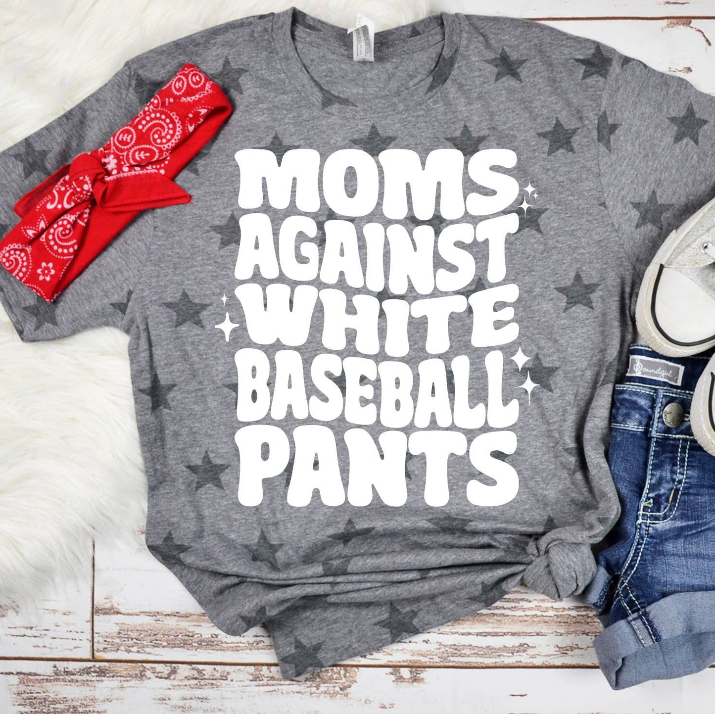 A flat-lay photo of the same gray t-shirt with the "MOMS AGAINST WHITE BASEBALL PANTS" design, paired with a red bandana and casual sneakers. A great outfit option for a sporty, relaxed look.