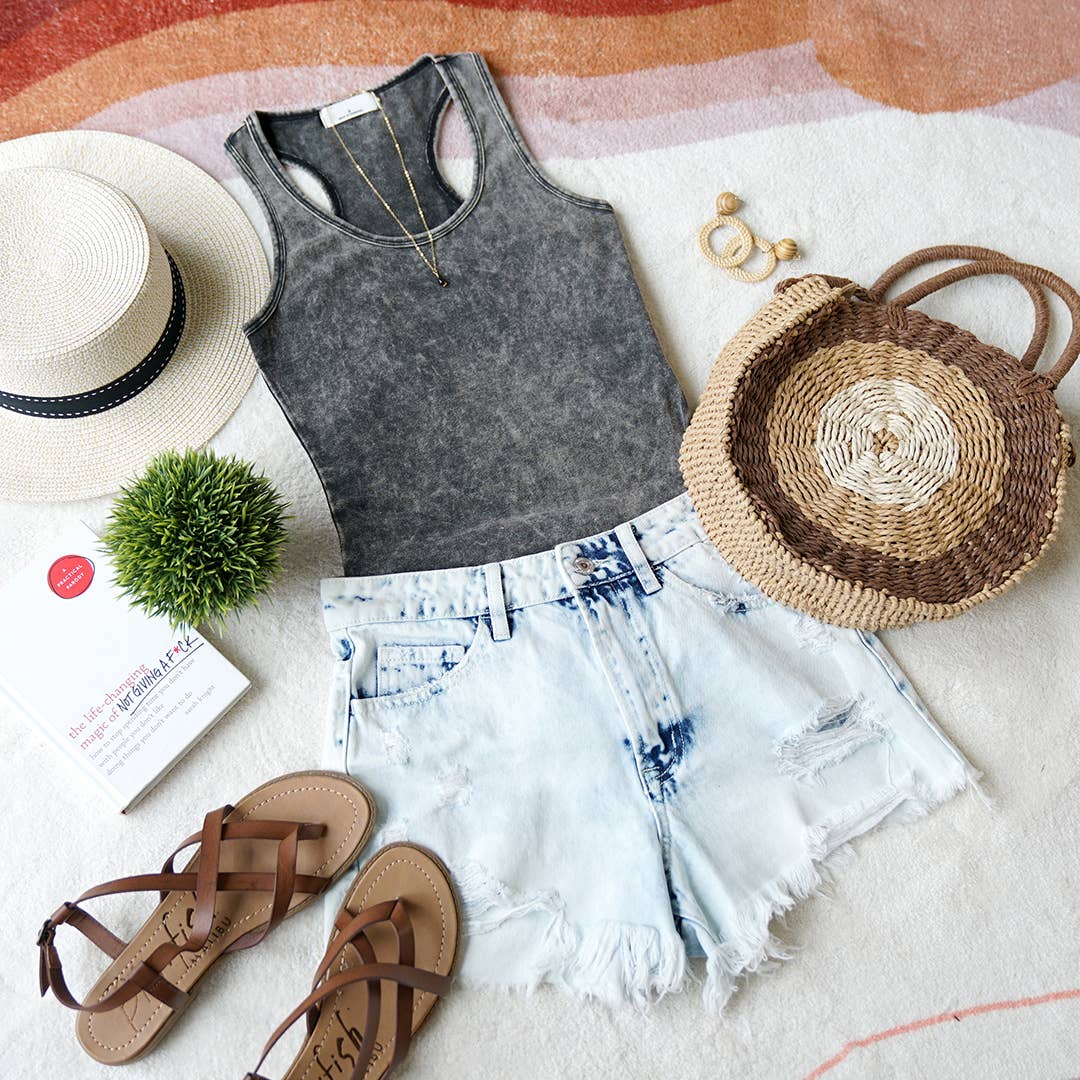Flat lay of a boho summer outfit with a gray acid-wash tank top, light blue distressed denim shorts, a straw bag, a straw hat, and other tank tops in earthy tones arranged alongside.