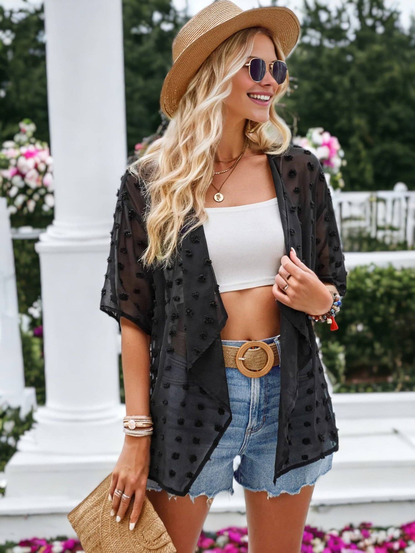 A stylish woman enjoying an outdoor setting wears a trendy summer outfit. She has on a sheer black kimono with textured polka dots over a white crop top, paired with denim shorts and a wide tan belt featuring a circular buckle. Accessories include a straw hat, oversized round sunglasses, layered gold necklaces, a woven clutch with a tassel, and colorful beaded bracelets. In the background, there are white pillars and blooming pink flowers, evoking a summery garden vibe.