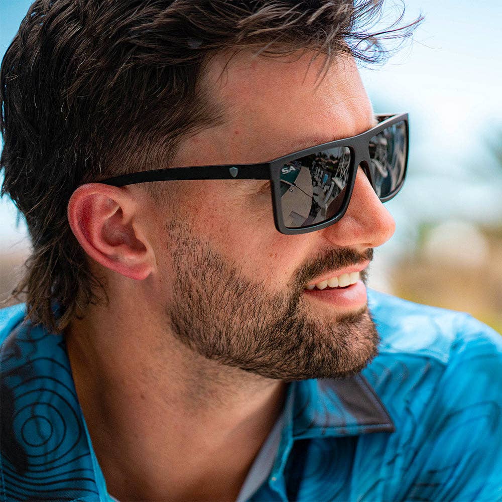 A man with short, dark hair and a beard wearing sleek black rectangular sunglasses. He is dressed in a blue patterned shirt, smiling while outdoors with a blurred background of sky and greenery.