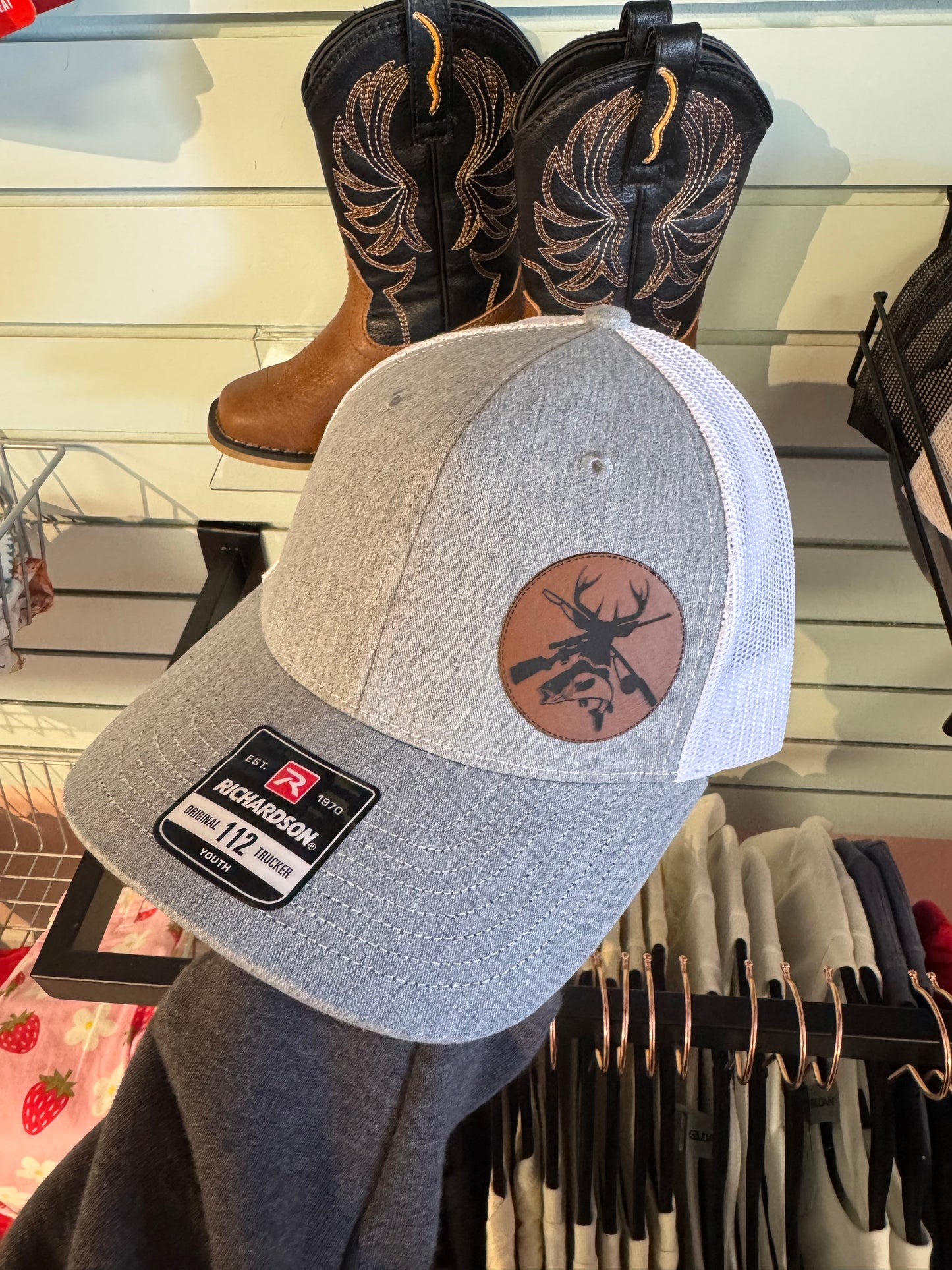 A gray and white trucker hat with a white mesh back and a circular brown leather patch on the front featuring a deer silhouette with crossed rifles. The hat is displayed on a retail rack with cowboy boots and clothing in the background.