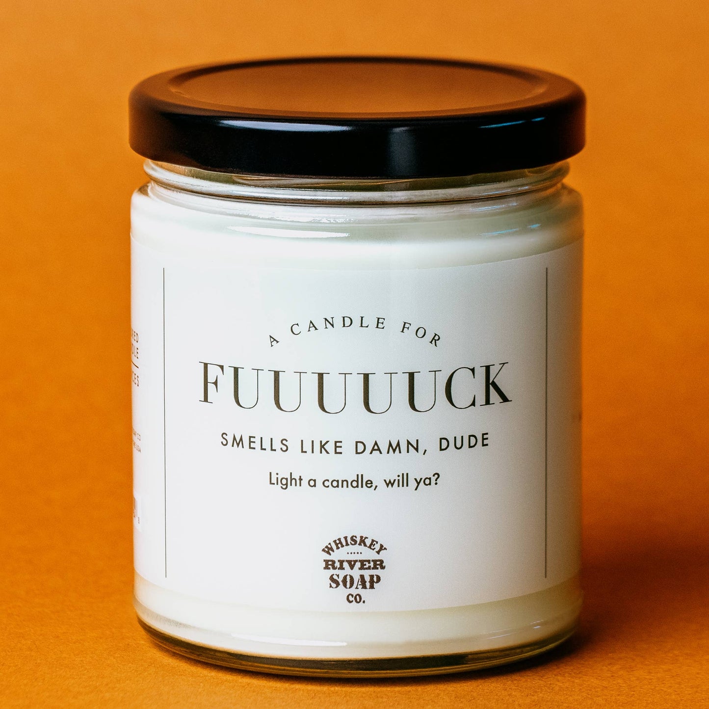 A humorous white candle in a clear glass jar with a black lid, labeled "A Candle For Fuuuuuuck" by Whiskey River Soap Co. The label humorously reads, "Smells like damn, dude. Light a candle, will ya?" Set against a warm orange background, this quirky product is perfect for anyone with a sense of humor and a need for some stress relief.