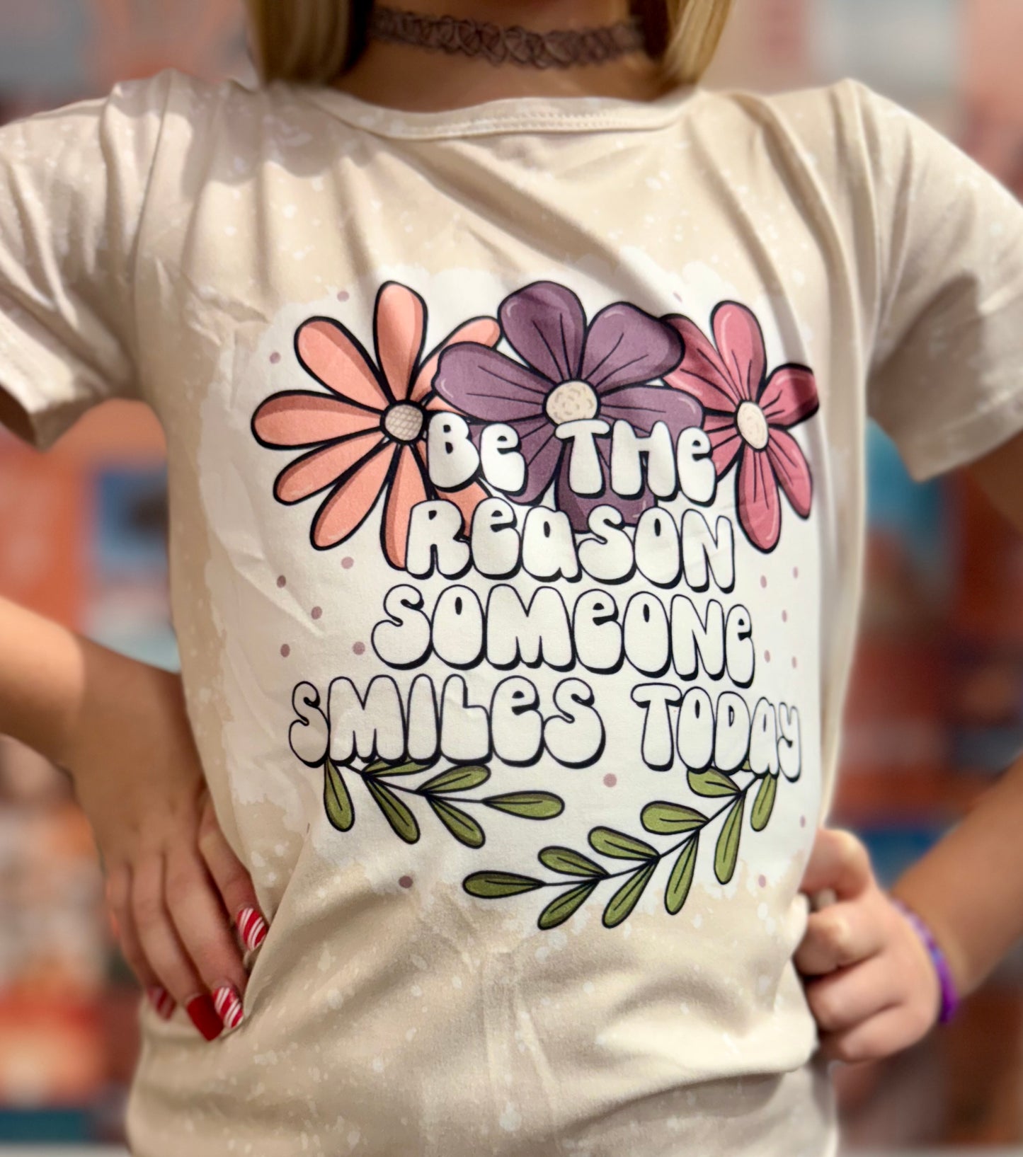 Beige short-sleeve T-shirt with a splatter design, featuring colorful floral graphics and the motivational phrase 'Be the reason someone smiles today' in bold, playful font. Paired with matching floral-print bell-bottom pants, this cheerful outfit is displayed on a pink hanger in a boutique setting, surrounded by vibrant accessories and other clothing items.