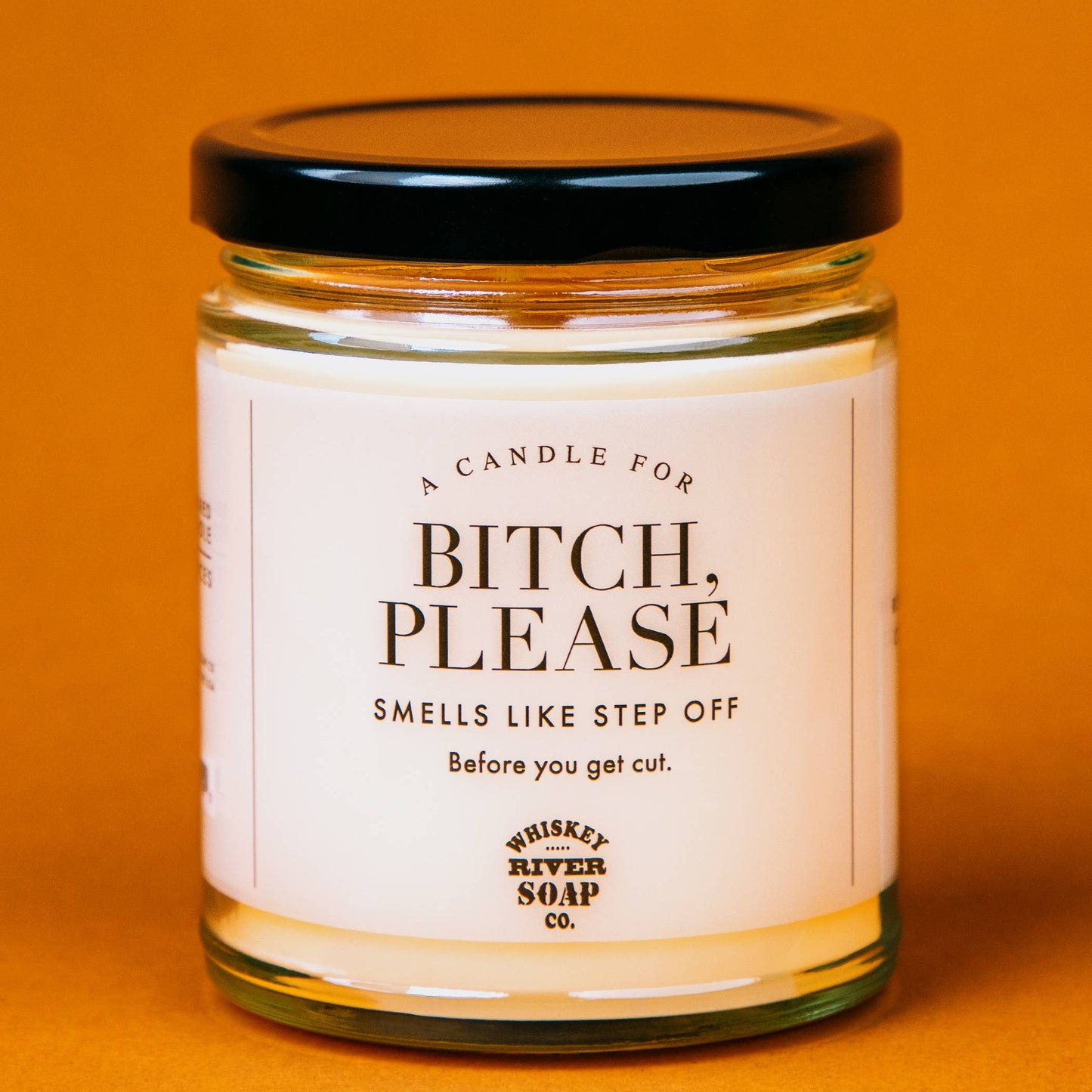 Humorous candle by Whiskey River Soap Co. labeled 'A Candle for Bitch, Please,' with the tagline 'Smells like step off before you get cut.' The design features a minimalist white jar with a black lid, set against a warm orange background, perfect for gifting with a touch of sass and personality.