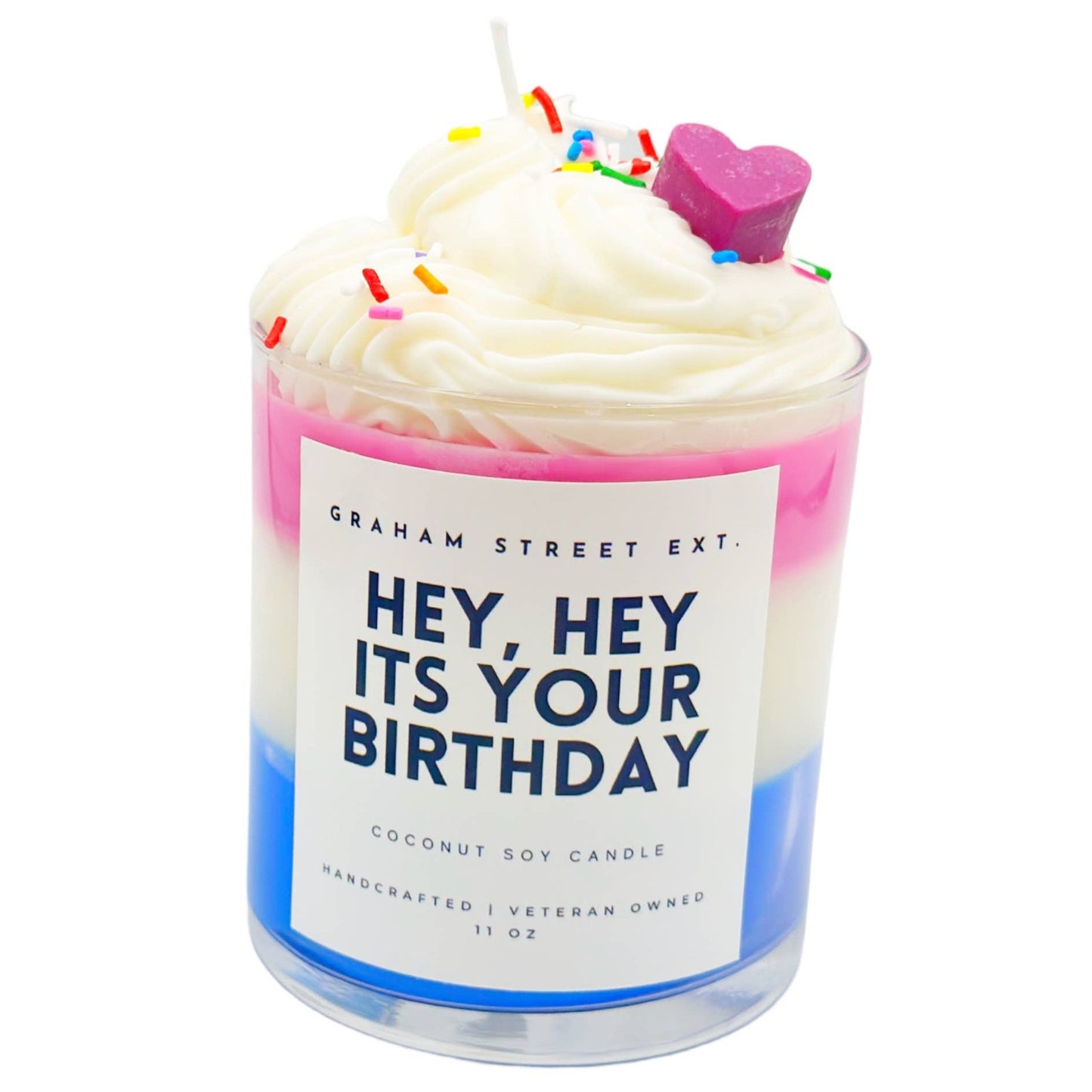 Festive coconut soy candle by Graham Street Ext., designed to resemble a birthday treat with layered pink, white, and blue wax. Topped with whipped cream-like frosting, colorful sprinkles, and a purple heart-shaped decoration, the label reads 'Hey, Hey It's Your Birthday.' This handcrafted, veteran-owned 11 oz candle adds a fun, celebratory touch to any occasion.