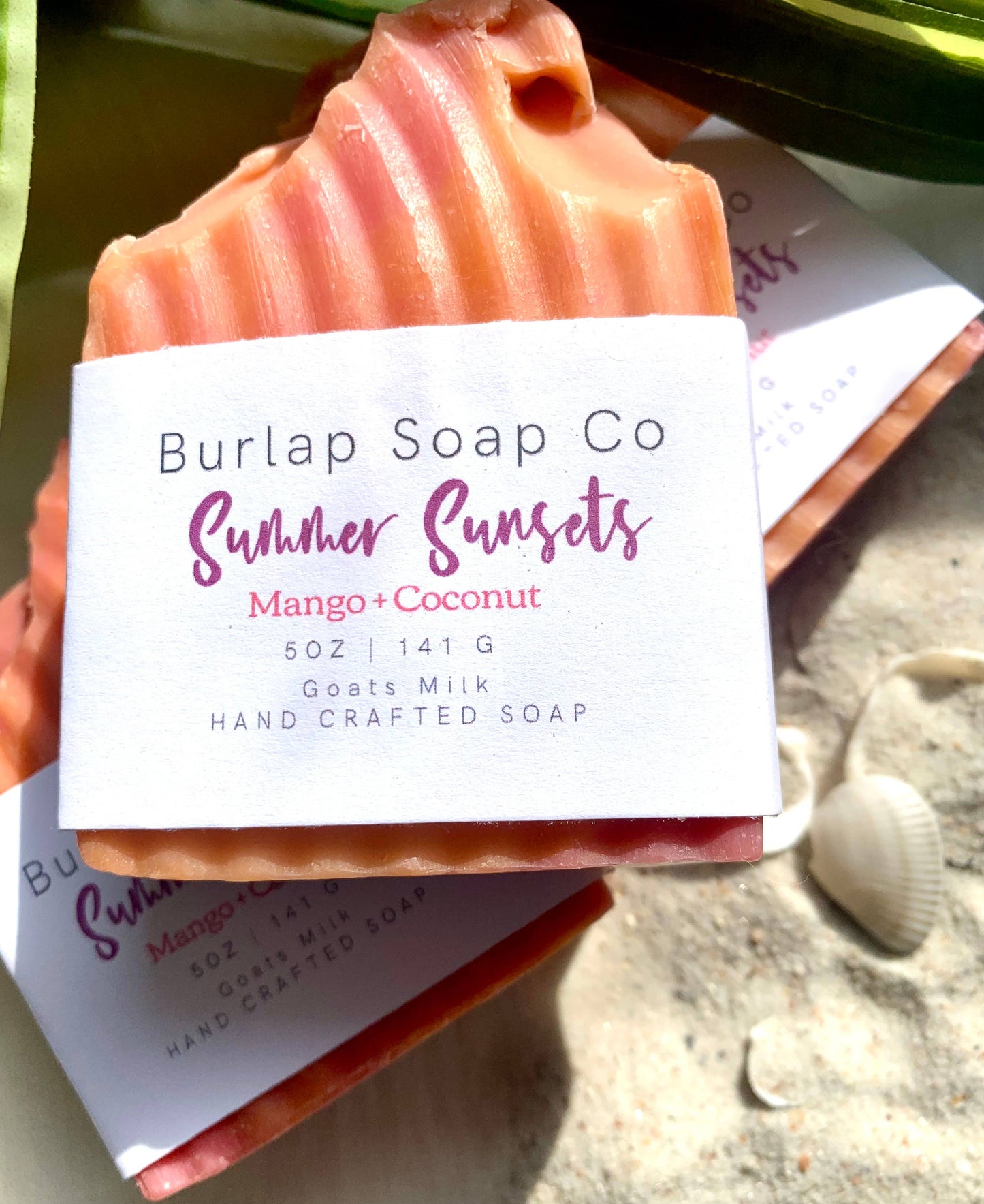 Close-up of Burlap Soap Co.'s handcrafted soap labeled 'Summer Sunsets Mango + Coconut,' made with goat's milk. The soap features vibrant orange and pink swirls, mimicking a tropical sunset, with its textured ridges. The product is displayed on a sandy surface with small seashells and green leaves, evoking a summery beach vibe.