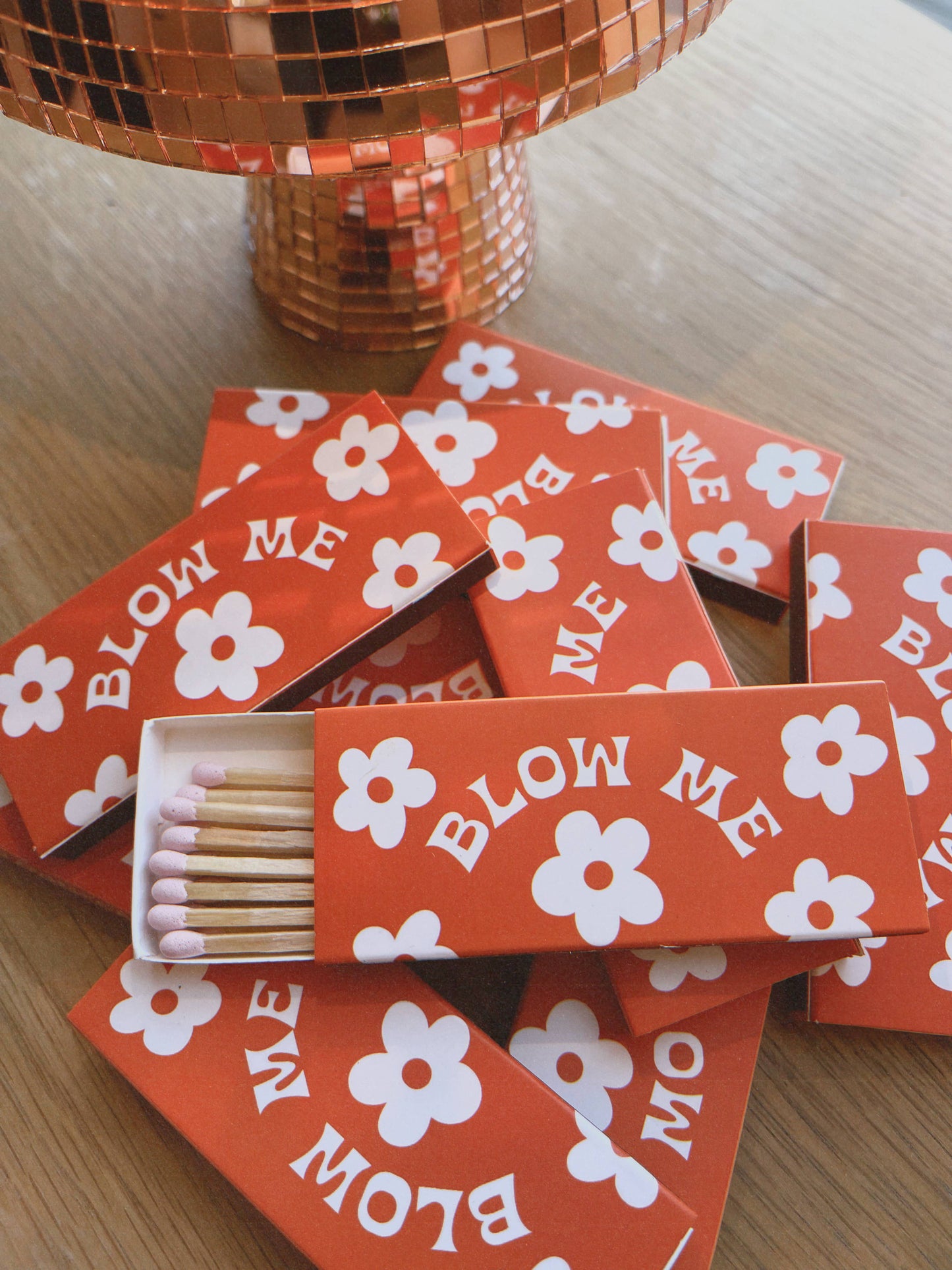 Stack of retro-inspired matchboxes with bright red covers featuring white daisy graphics and bold text reading "BLOW ME." One box is open, revealing matches with pink tips. Displayed on a wooden surface beside a copper disco-ball-style decor piece, adding a playful, vintage vibe.